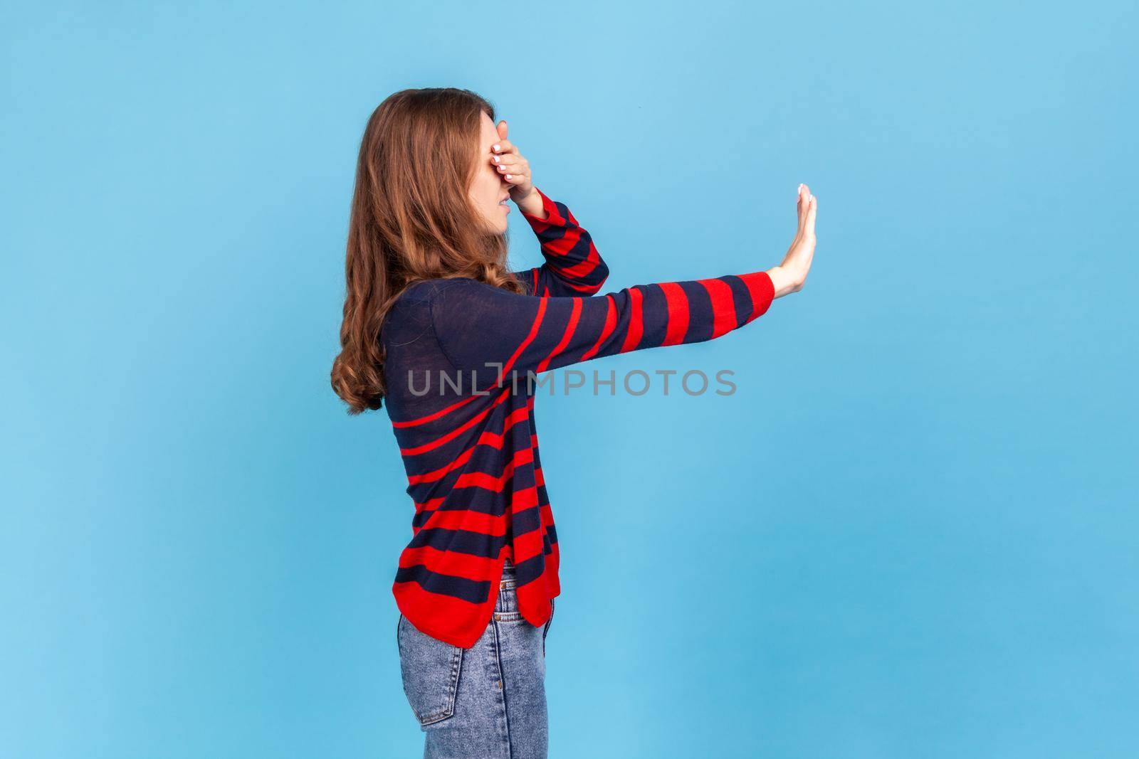 Side view portrait of woman wearing striped casual style sweater standing and afraid, covering her eyes with hand and trying to blocking Indoor studio shot isolated on blue background.
