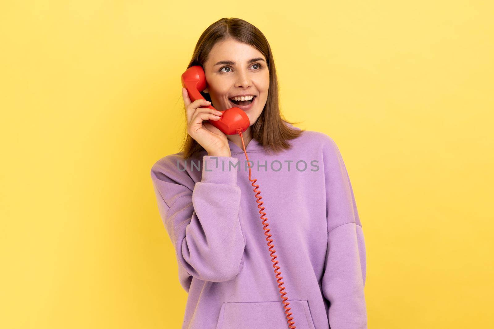 Portrait of beautiful young adult woman with excited expression talking on red retro telephone, holding handset, wearing purple hoodie. Indoor studio shot isolated on yellow background.