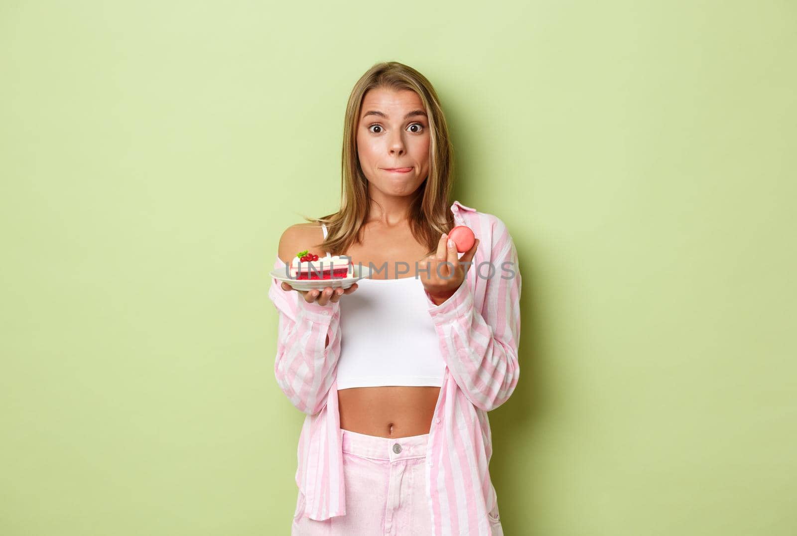 Image of attractive blond woman looking tempted and licking lips, holding macaroons and tasty cake, standing over green background.