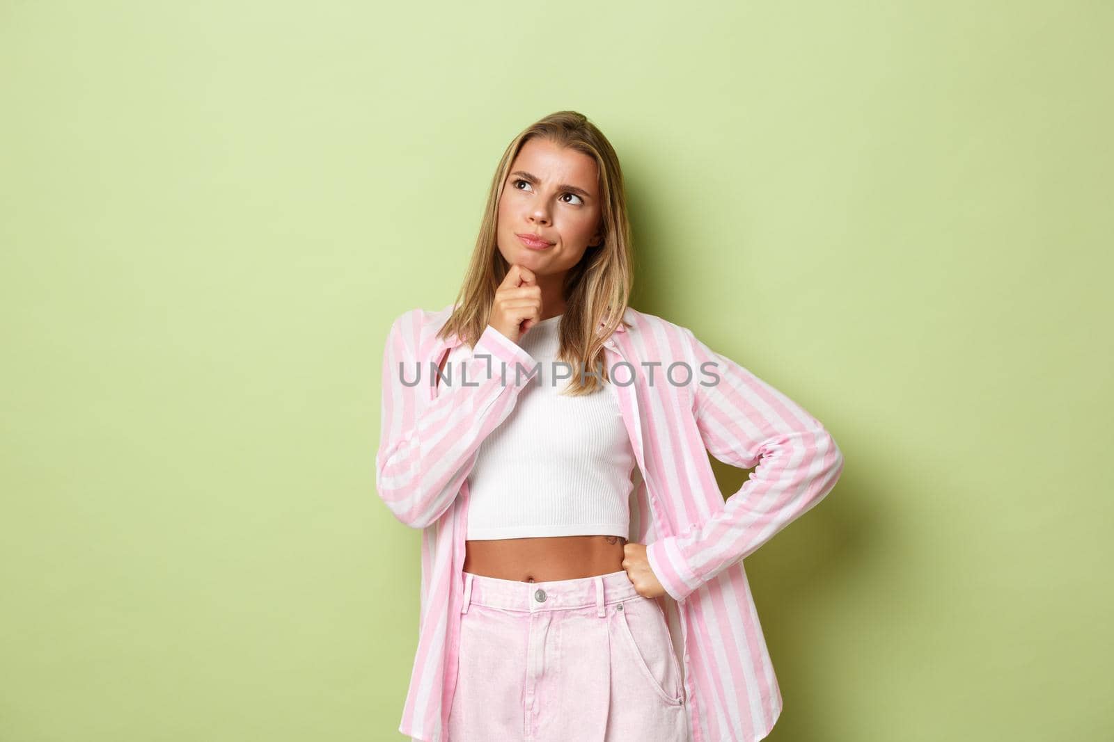 Image of thoughtful young female in pink outfit, looking at upper left corner and thinking, making difficult choice, standing over green background.