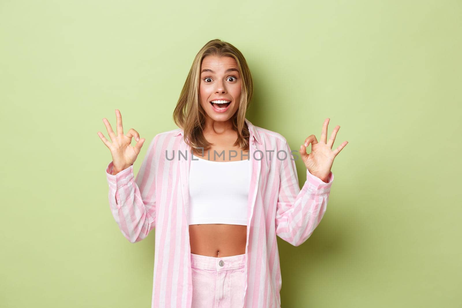 Portrait of attractive stylish woman with blond short hairstyle, wearing pink shirt with jeans, showing okay sign in approval, recommend something good, standing over green background.