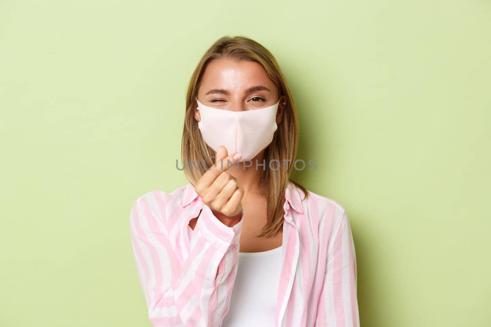 Concept of quarantine, coronavirus and lifestyle. Close-up of beautiful blond female model in face mask and pink shirt, showing heart sign, standing over green background by Benzoix