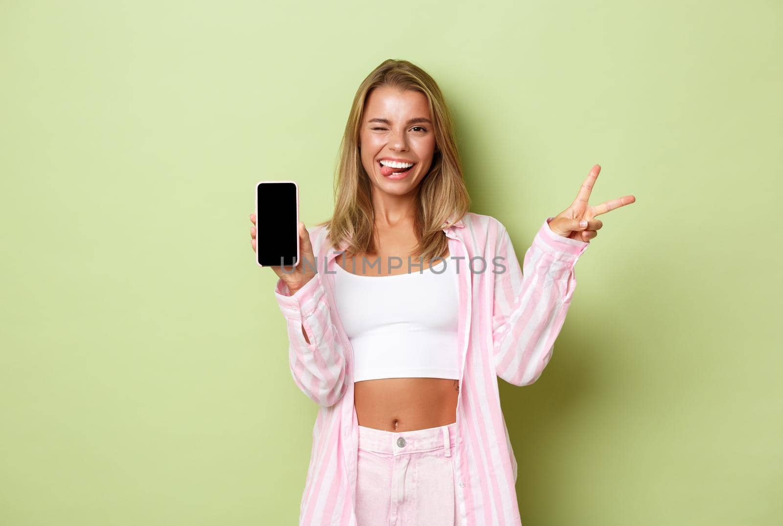 Portrait of cute blond girl in pink shirt, showing peace sign and mobile phone screen, smiling pleased, standing over green background by Benzoix