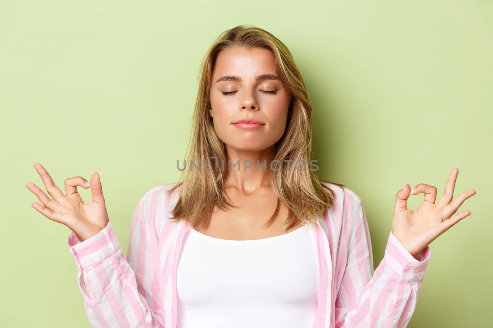 Close-up of peaceful blond girl in pink shirt, meditating with closed eyes, practice yoga, standing patient over green background by Benzoix