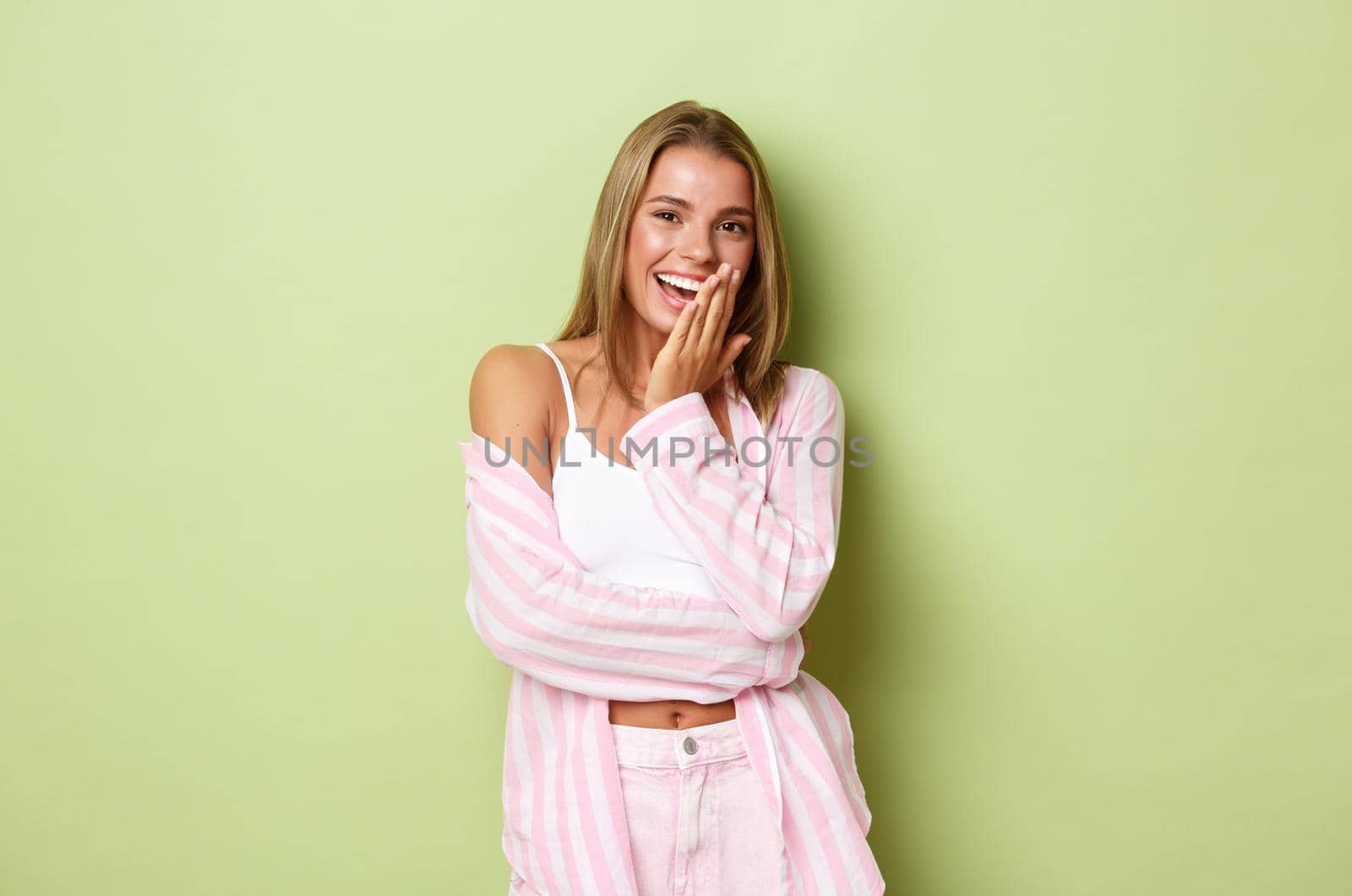 Portrait of flirty blond woman in pink shirt, laughing and cover mouth coquettish, smiling happy over something funny, standing over green background by Benzoix