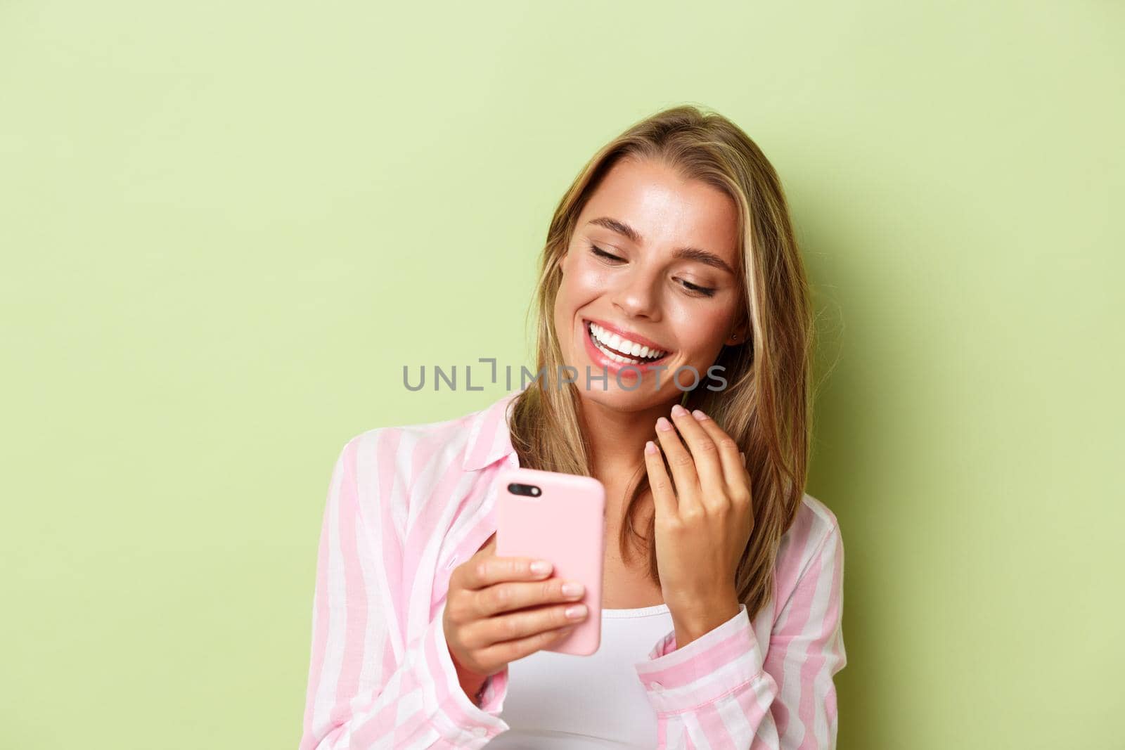 Close-up of attractive blond girl in pink shirt, messaging on phone, smiling and looking at smartphone, standing over green background.