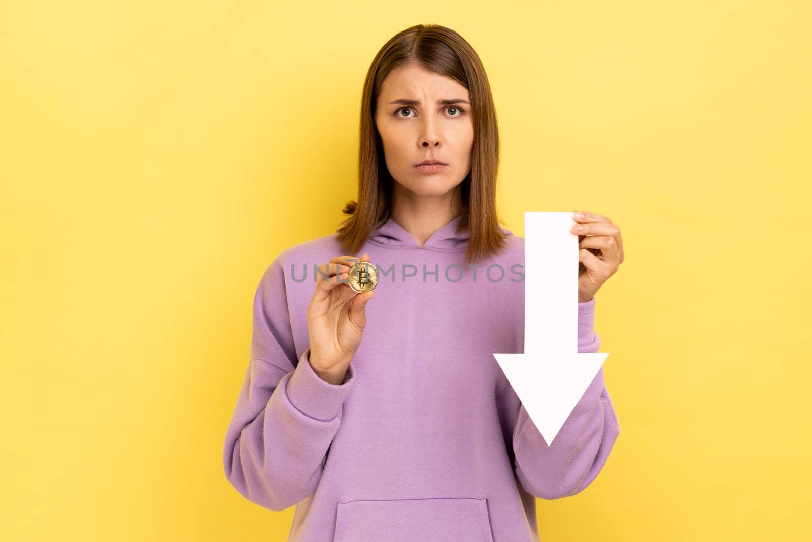 Portrait of unhappy upset woman showing bitcoin and white arrow pointing down, expressing negative emotions, wearing purple hoodie. Indoor studio shot isolated on yellow background.