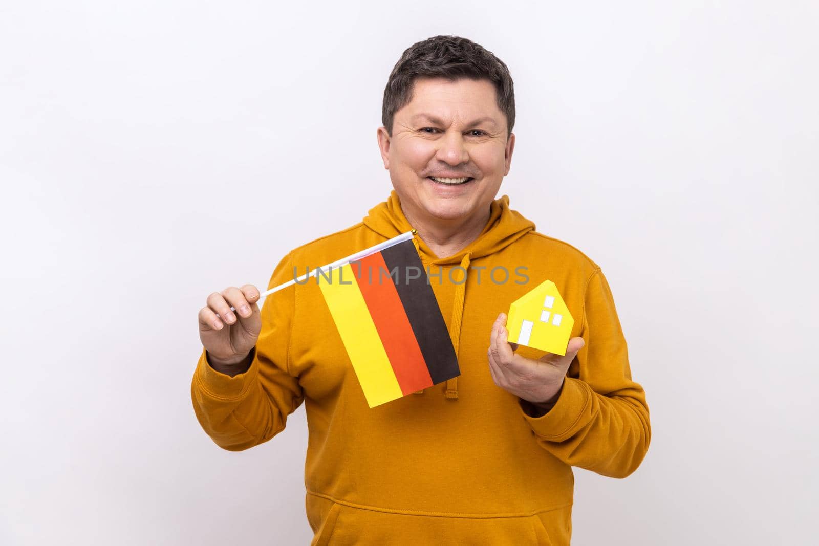 Optimistic man holding German flag and paper house, dreaming to buy his own apartrment in Germany, looking at camera, wearing urban style hoodie. Indoor studio shot isolated on white background.