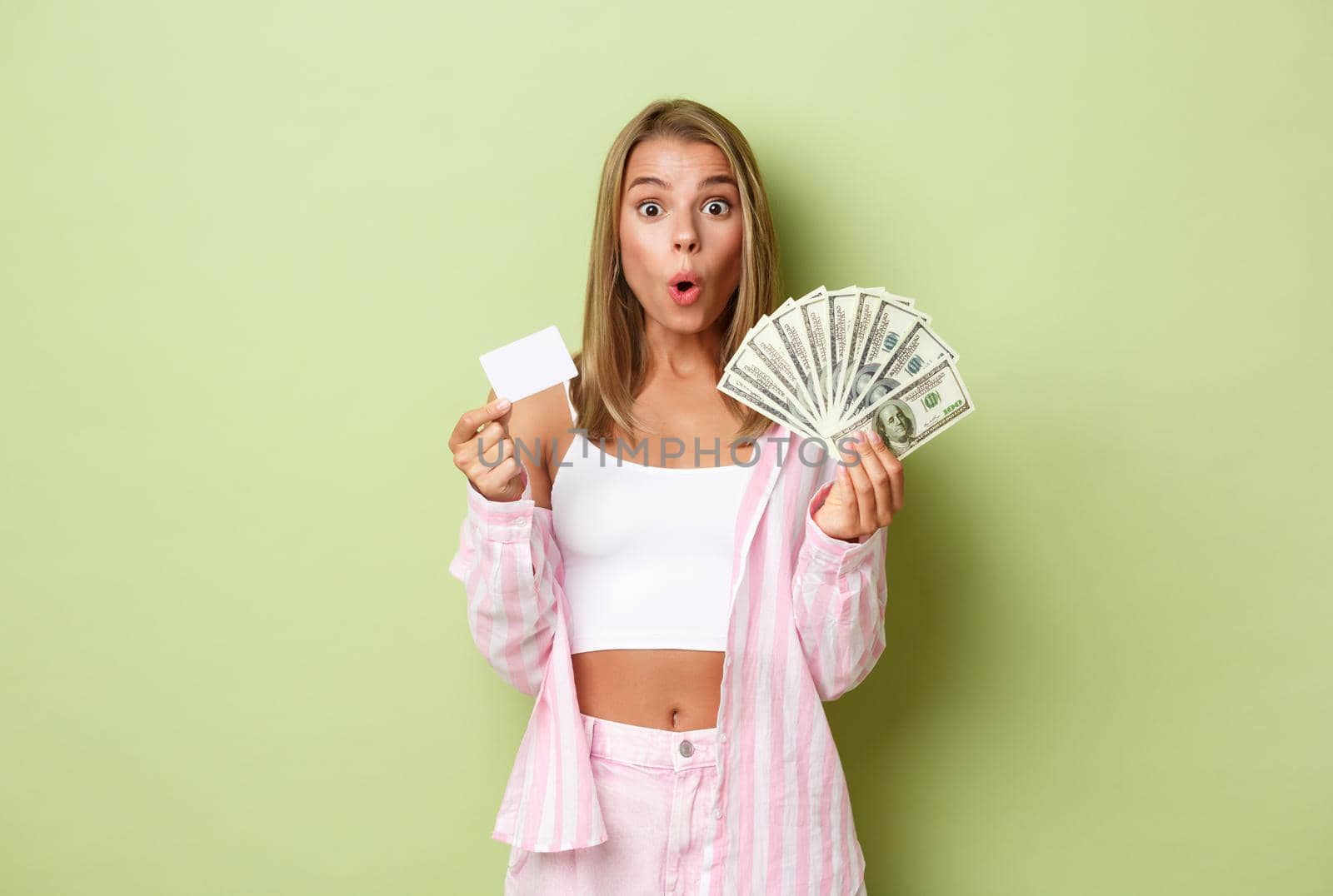 Image of impressed and excited blond woman, holding cash and credit card, standing over green background by Benzoix