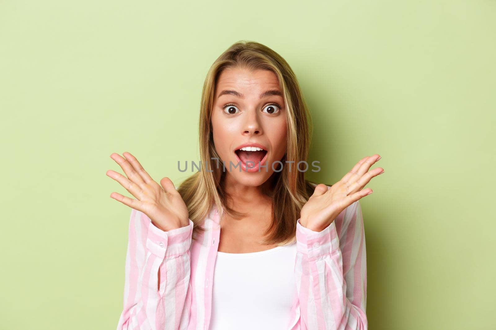 Close-up of attractive blond woman looking surprised, spread hands sideways and open mouth impressed, standing over green background.