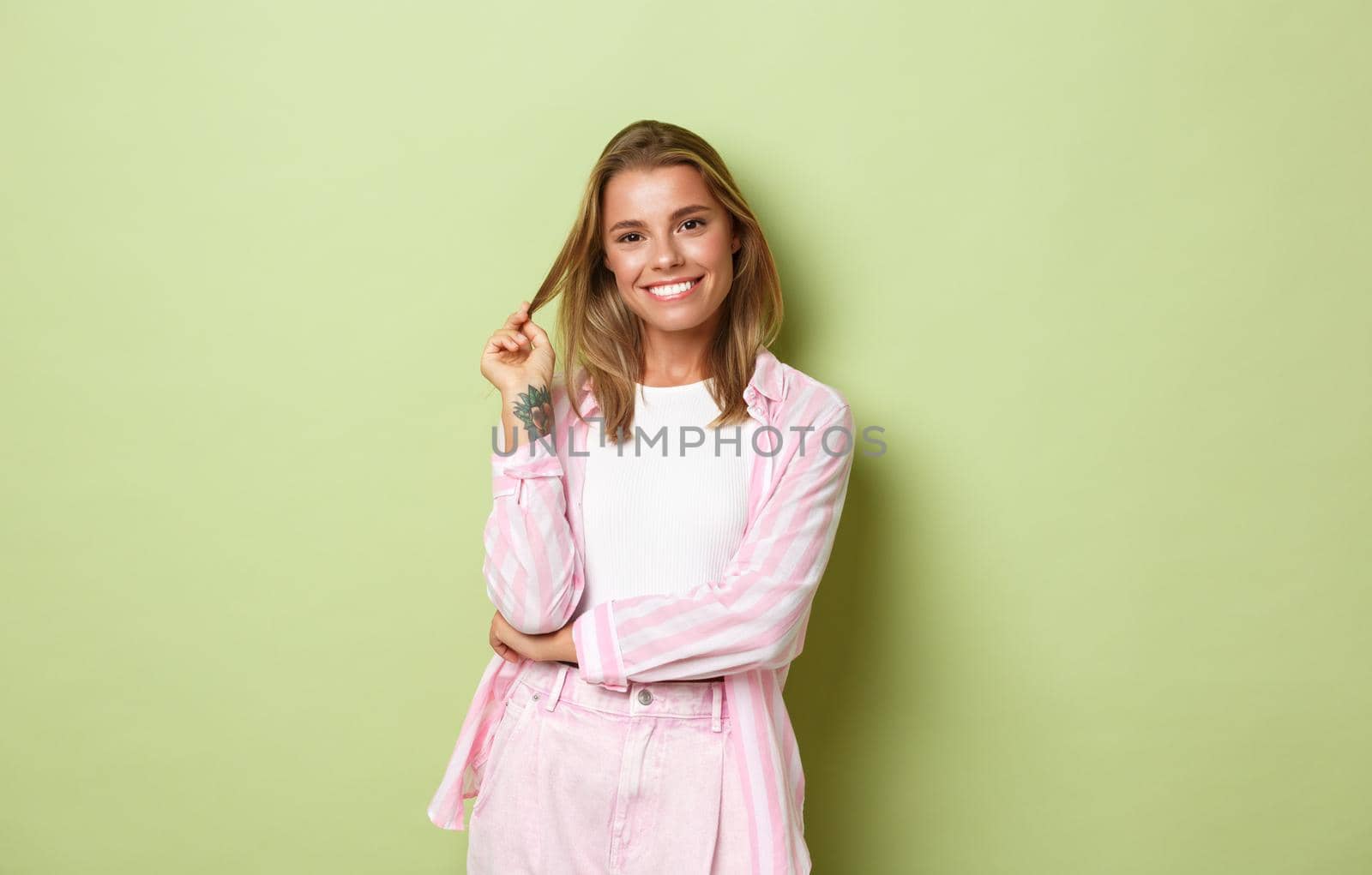 Image of beautiful caucasian woman with short blond hairstyle, tattoos, wearing pink costume, smiling confident and pleased, standing over green background by Benzoix