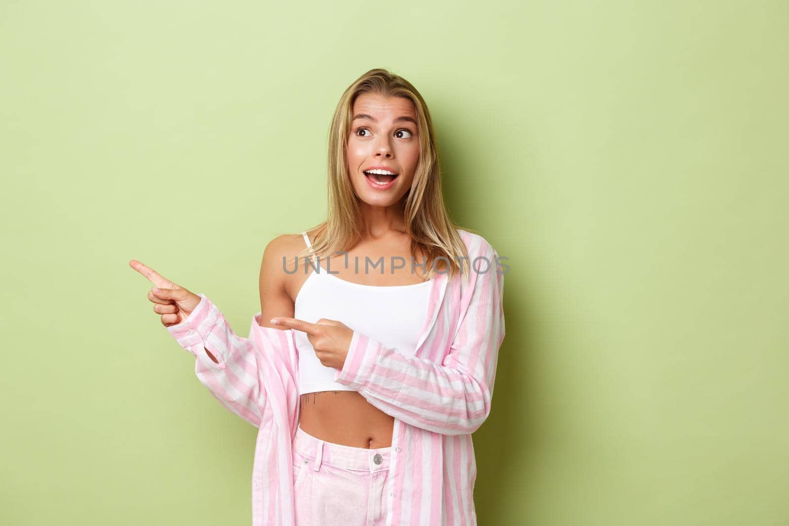 Attractive cheerful girl with short blond hairstyle, looking amused and pointing fingers left at advertising, standing over green background by Benzoix