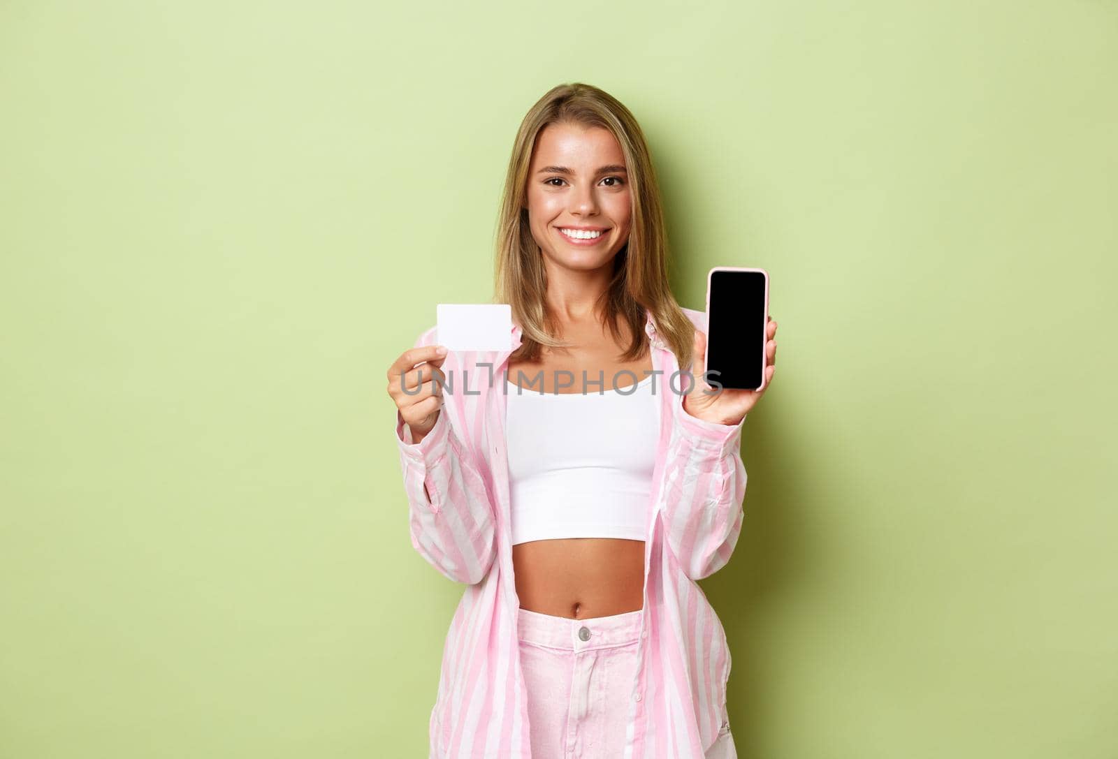 Image of stylish blond woman in pink shirt and jeans, showing smarpthone screen and credit card, recommending something online, standing over green background.