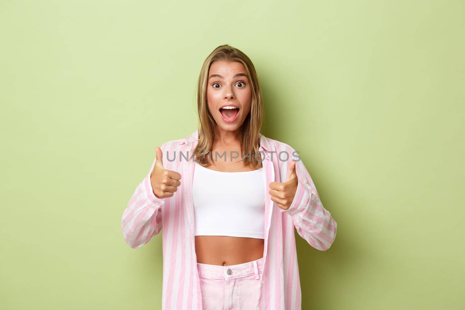 Image of surprised happy female model with blond hair, wearing pink shirt and jeans, showing thumbs-up in approval, like something, standing over green background by Benzoix