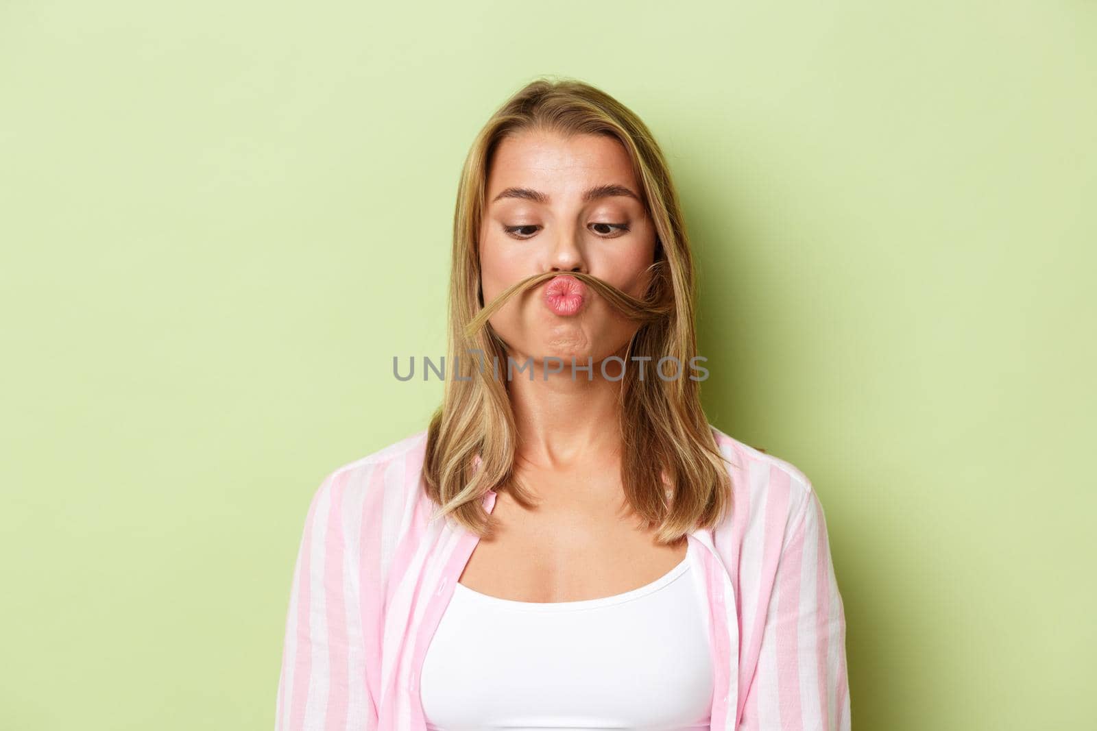 Close-up of attractive blond girl in pink shirt, making moustache with hair strand, playing and fooling around, standing over green background.