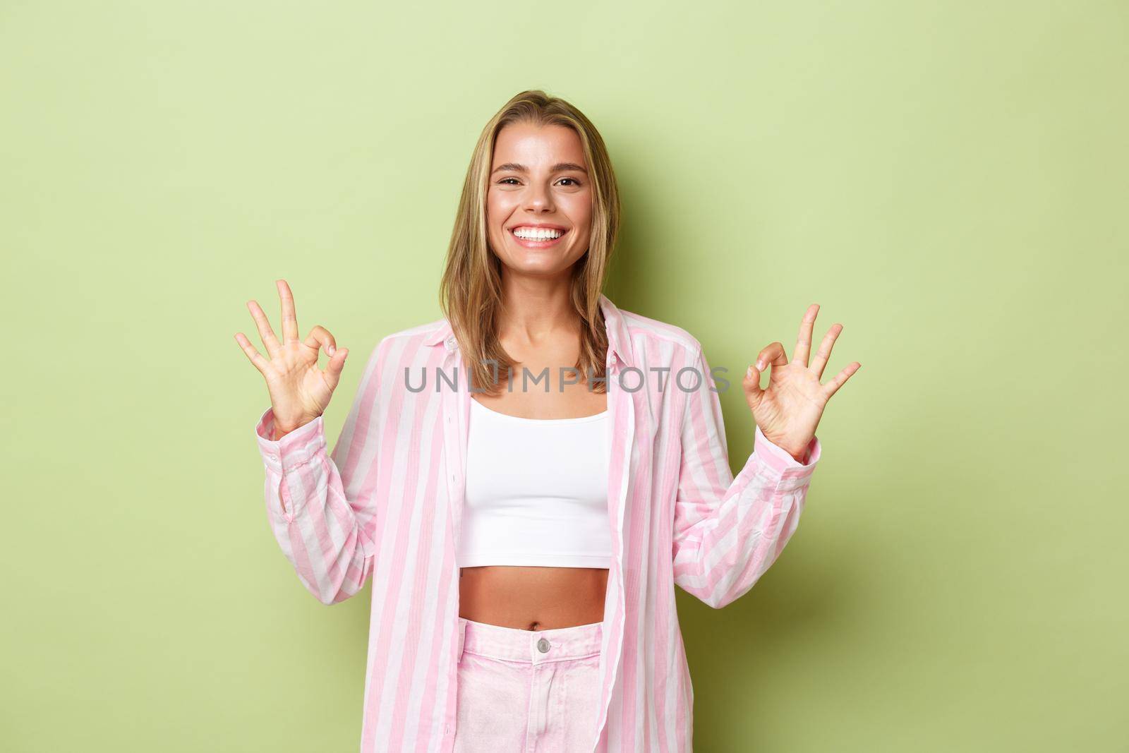 Portrait of attractive stylish woman with blond short hairstyle, wearing pink shirt with jeans, showing okay sign in approval, recommend something good, standing over green background.