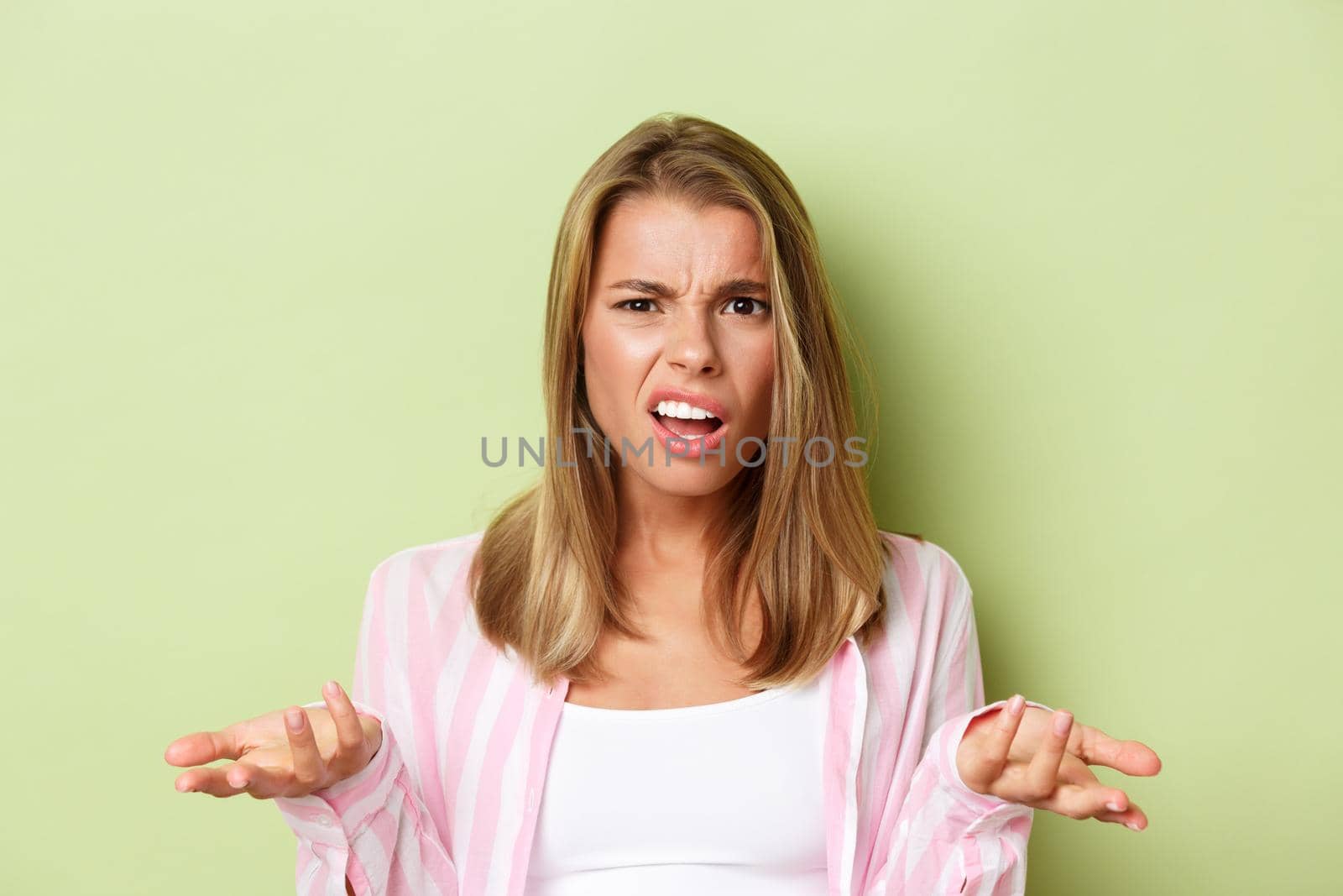 Close-up of pretty blond woman looking confused, raise hands sideways and arguing about something, standing over green background by Benzoix