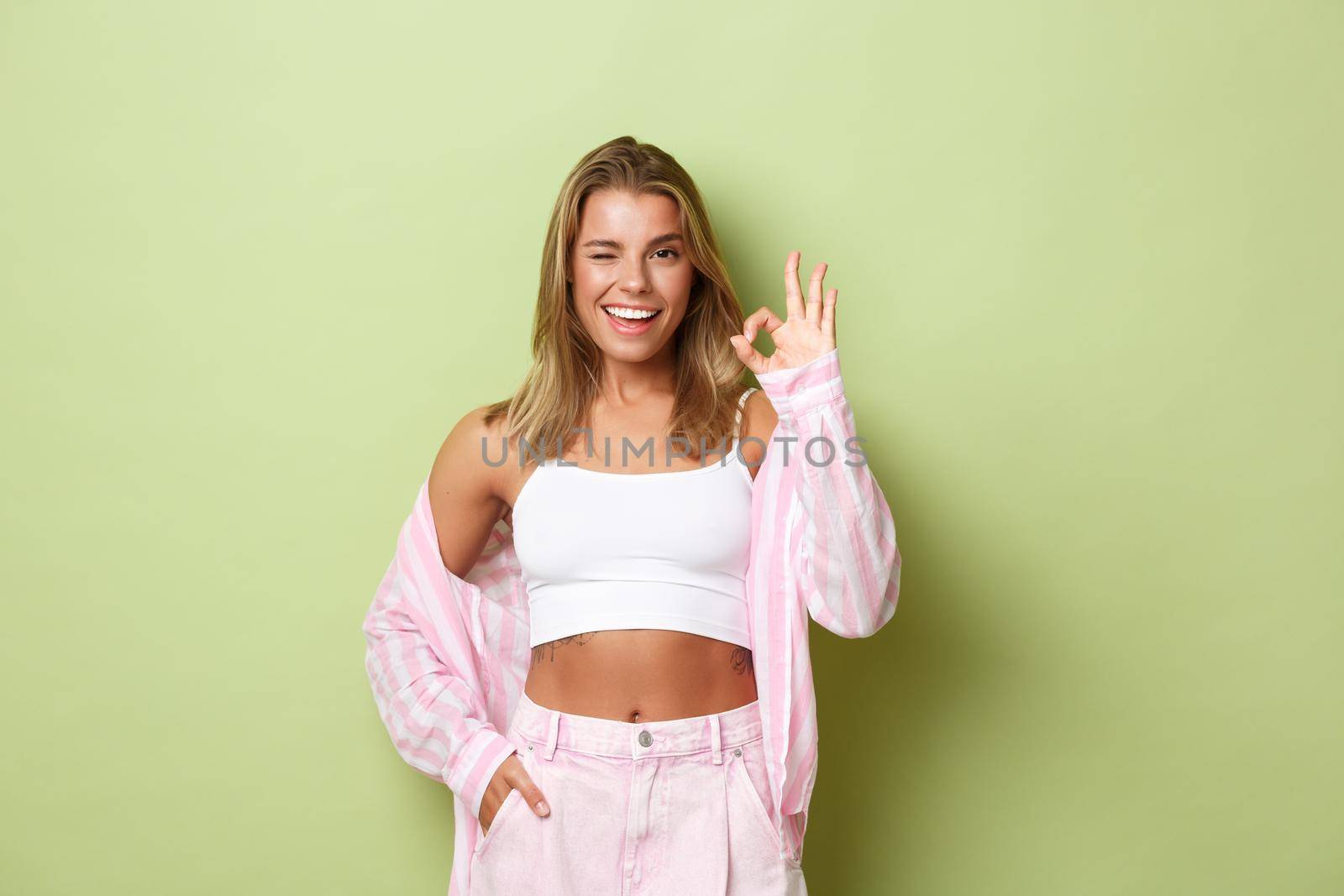 Portrait of stylish blond woman in pink costume and tank-top, showing okay sign and winking, guarantee and recommend something good, standing over green background by Benzoix