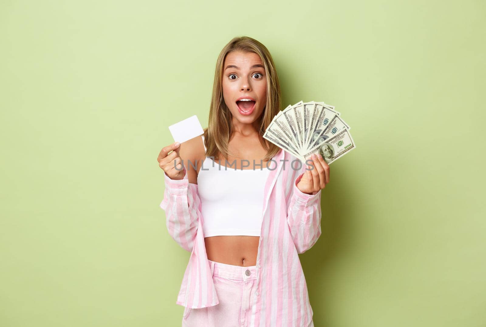 Portrait of excited blond woman showing credit card and cash, standing amazed over green background by Benzoix