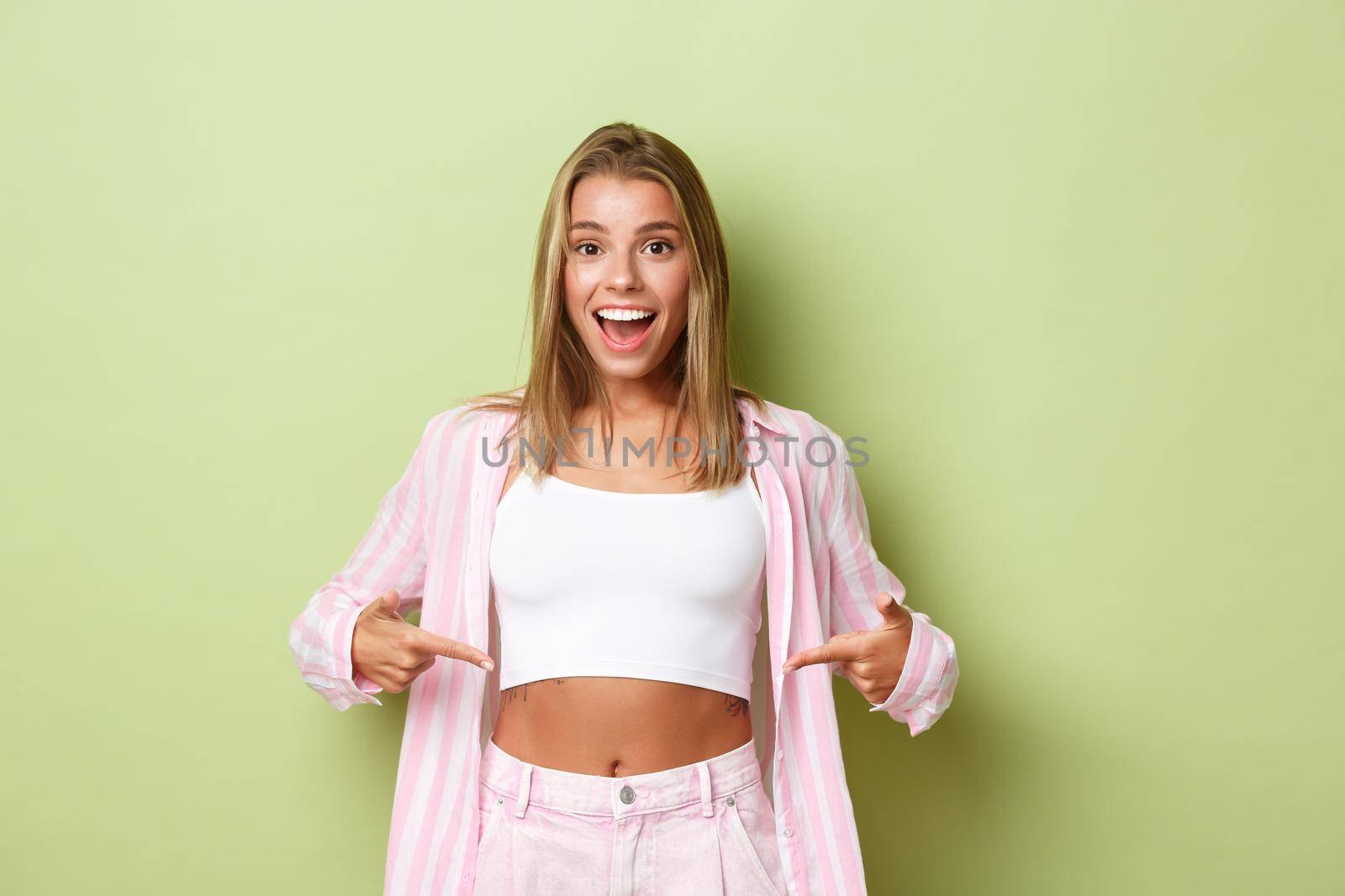 Image of happy blond woman with perfect body and white smile, pointing fingers at her flat belly, showing workout results, standing over green background.