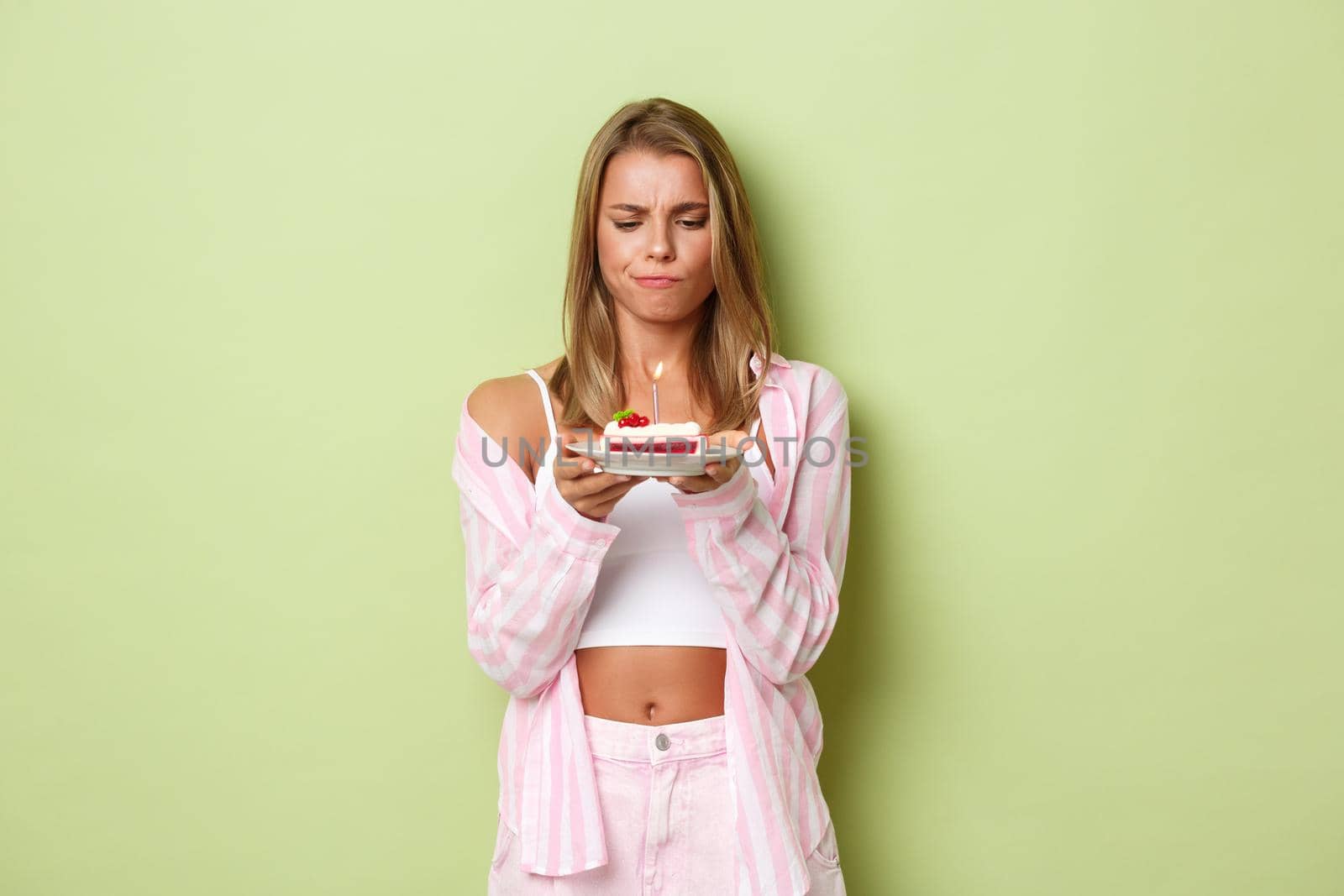 Image of indecisive blond girl pouting while looking at birthday cake with lit candle, thinking about bday wish, standing over green background.