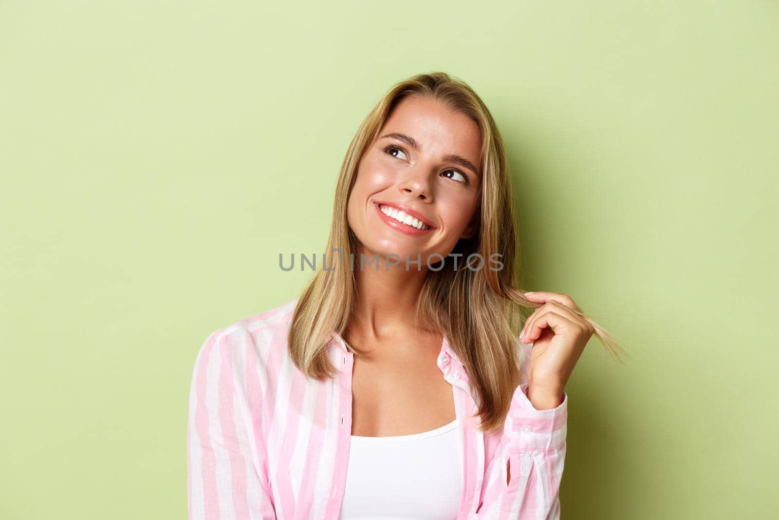 Close-up of glamour blond girl touching hair strand, smiling and looking dreamy at upper left corner, standing over green background by Benzoix