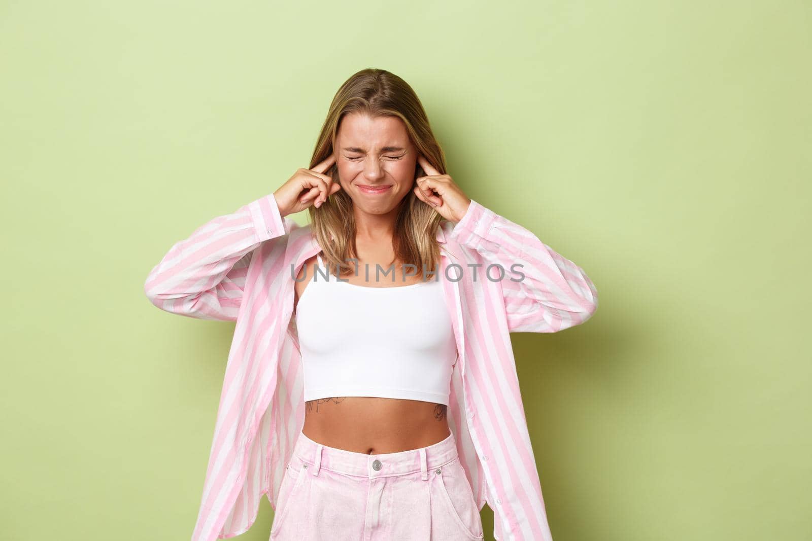 Portrait of annoyed blond woman feeling uncomfortable, shut ears with fingers from loud noise, standing bothered over green background by Benzoix