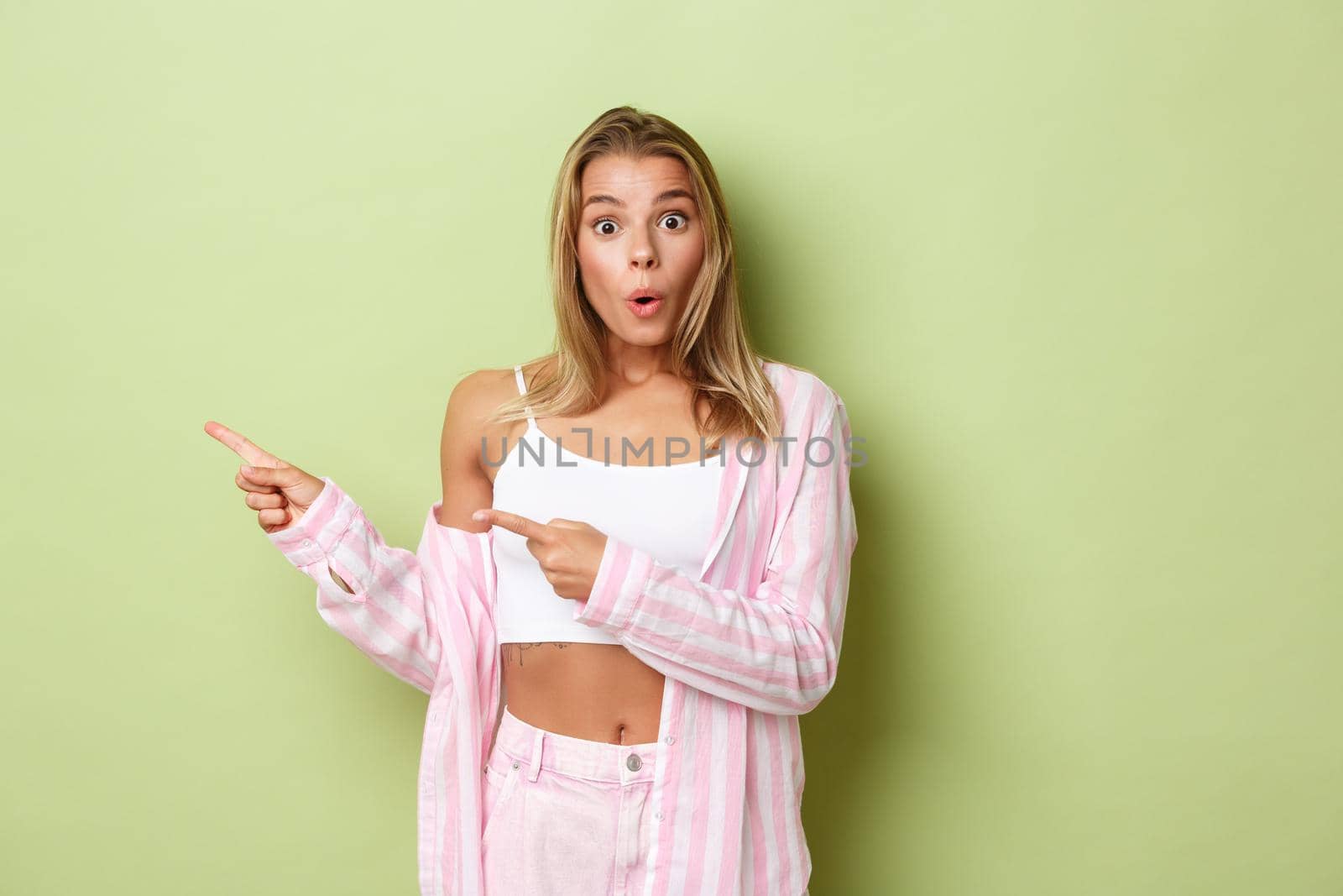 Surprised attractive blond girl in pink shirt and jeans pointing fingers left, showing your logo on green background.