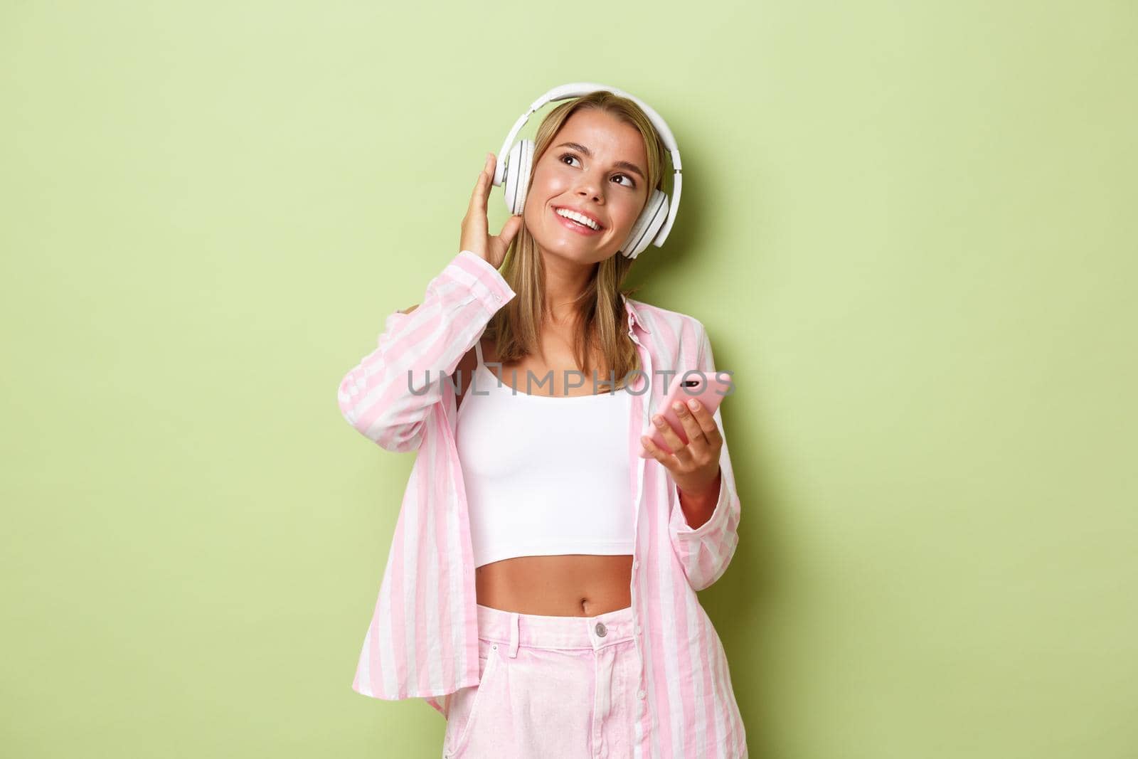 Image of stylish glamour girl with blond short hair, looking at upper left corner dreamy while listening to music in wireless headphones, holding mobile phone, green background.