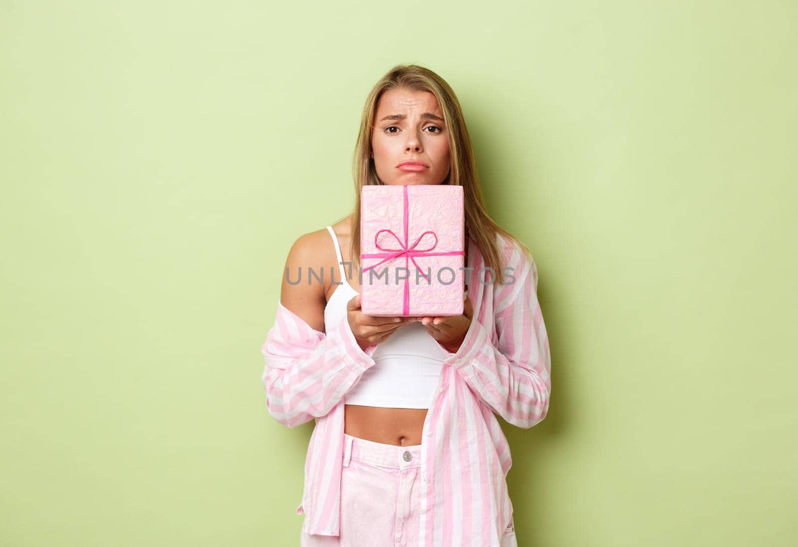 Image of gloomy blond woman sulking, holding pink gift and frowning, standing against green background disappointed.