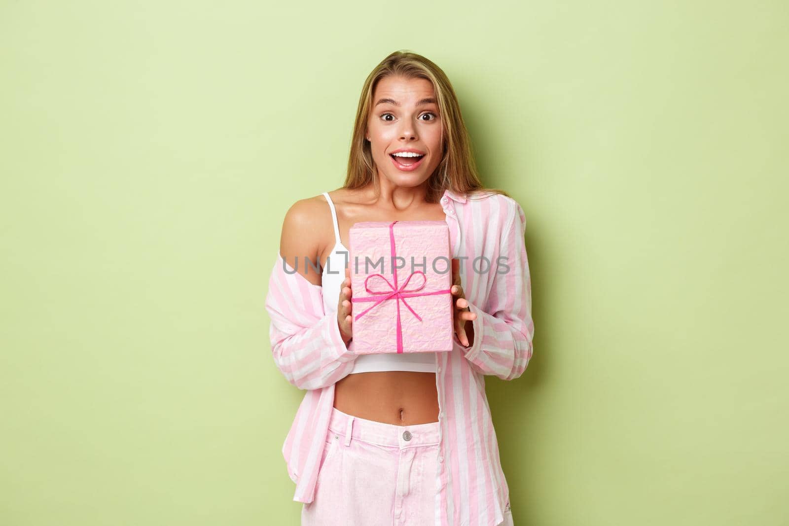 Portrait of attractive modern girl with blond short hair, looking excited, receiving a gift and smiling, standing over green background by Benzoix