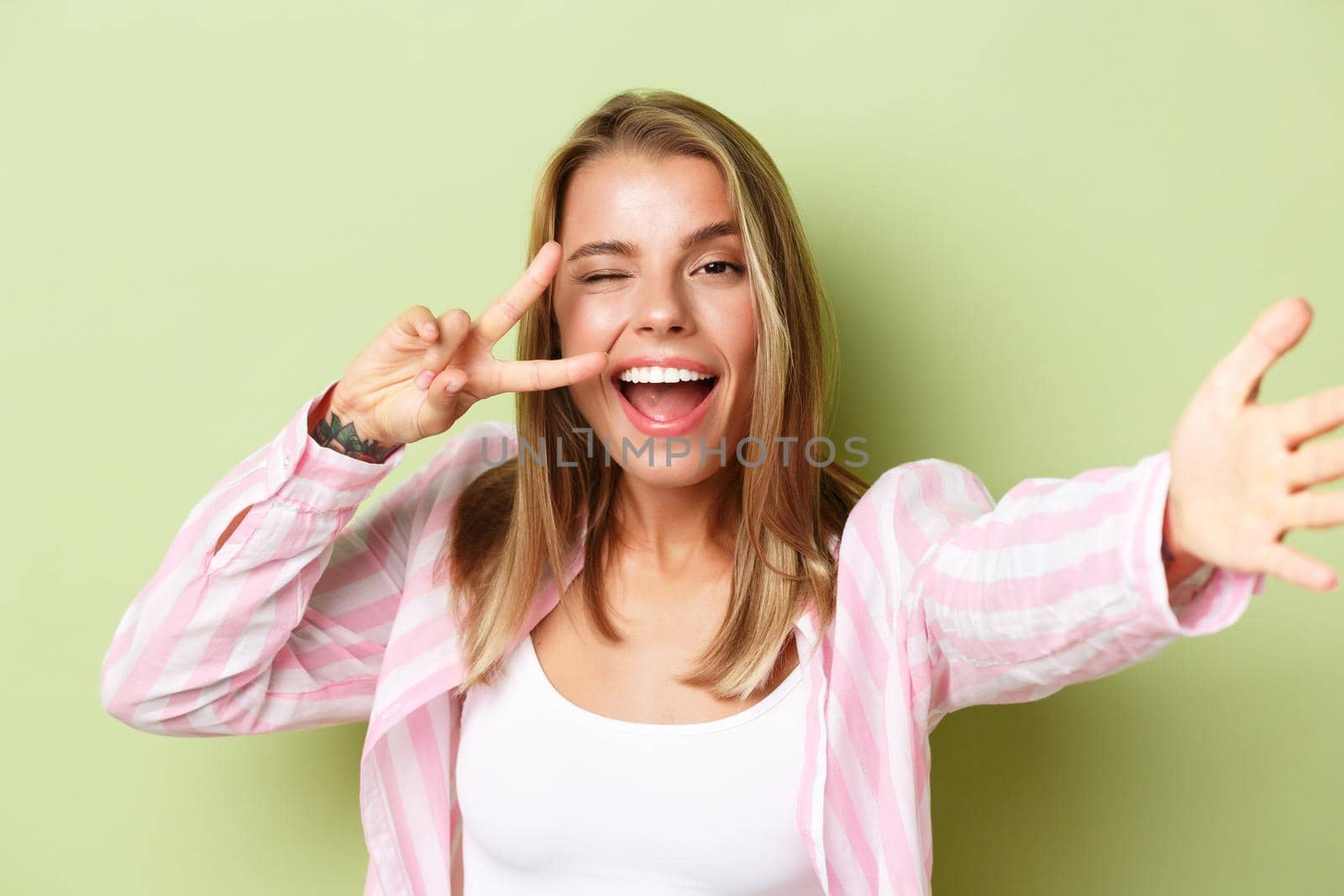 Close-up of stylish blond girl with tattoo, wearing pink shirt, taking selfie, showing peace sign at camera and smiling, standing over green background by Benzoix