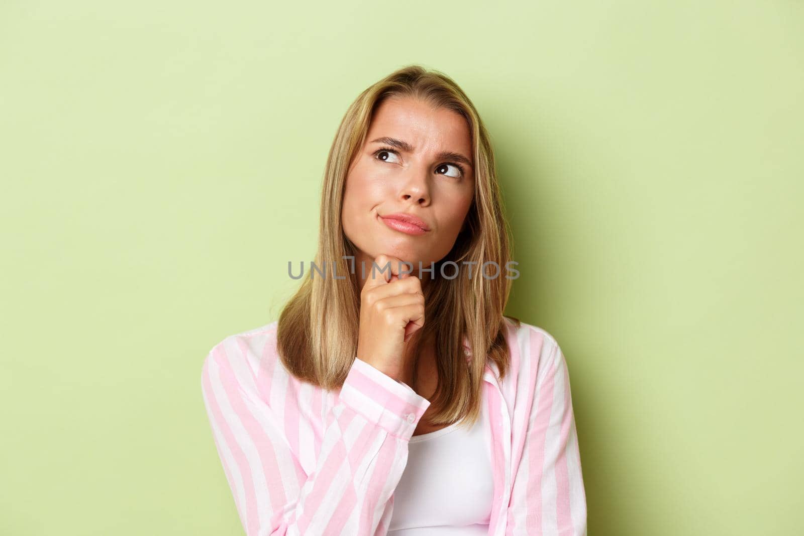 Close-up of thoughtful blond girl in pink shirt, touching chin and looking at upper left corner, making decision, green background.