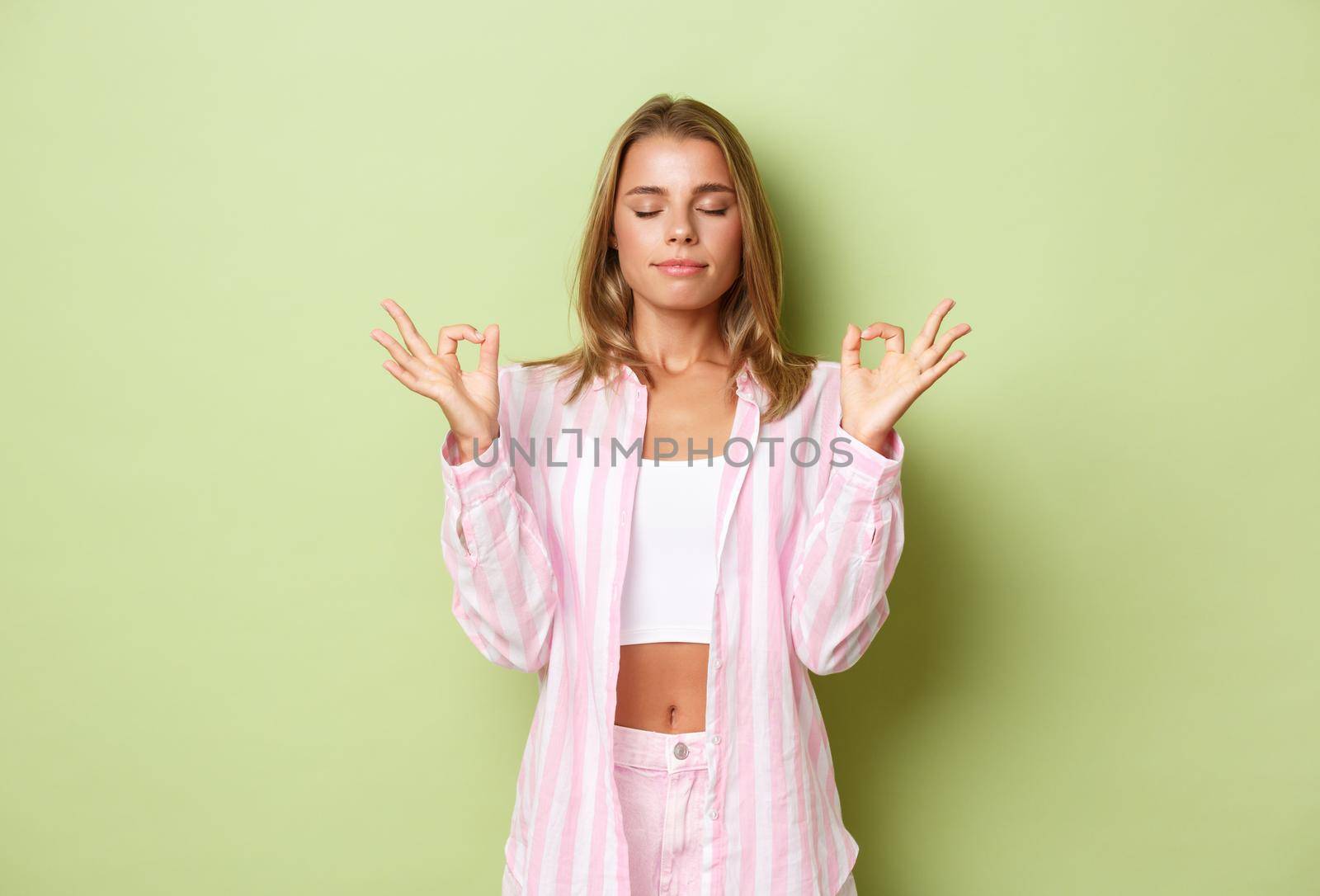 Portrait of young relaxed woman with blond short hairstyle, closing eyes and holding hands in zen gesture, meditating, practice yoga over green background.