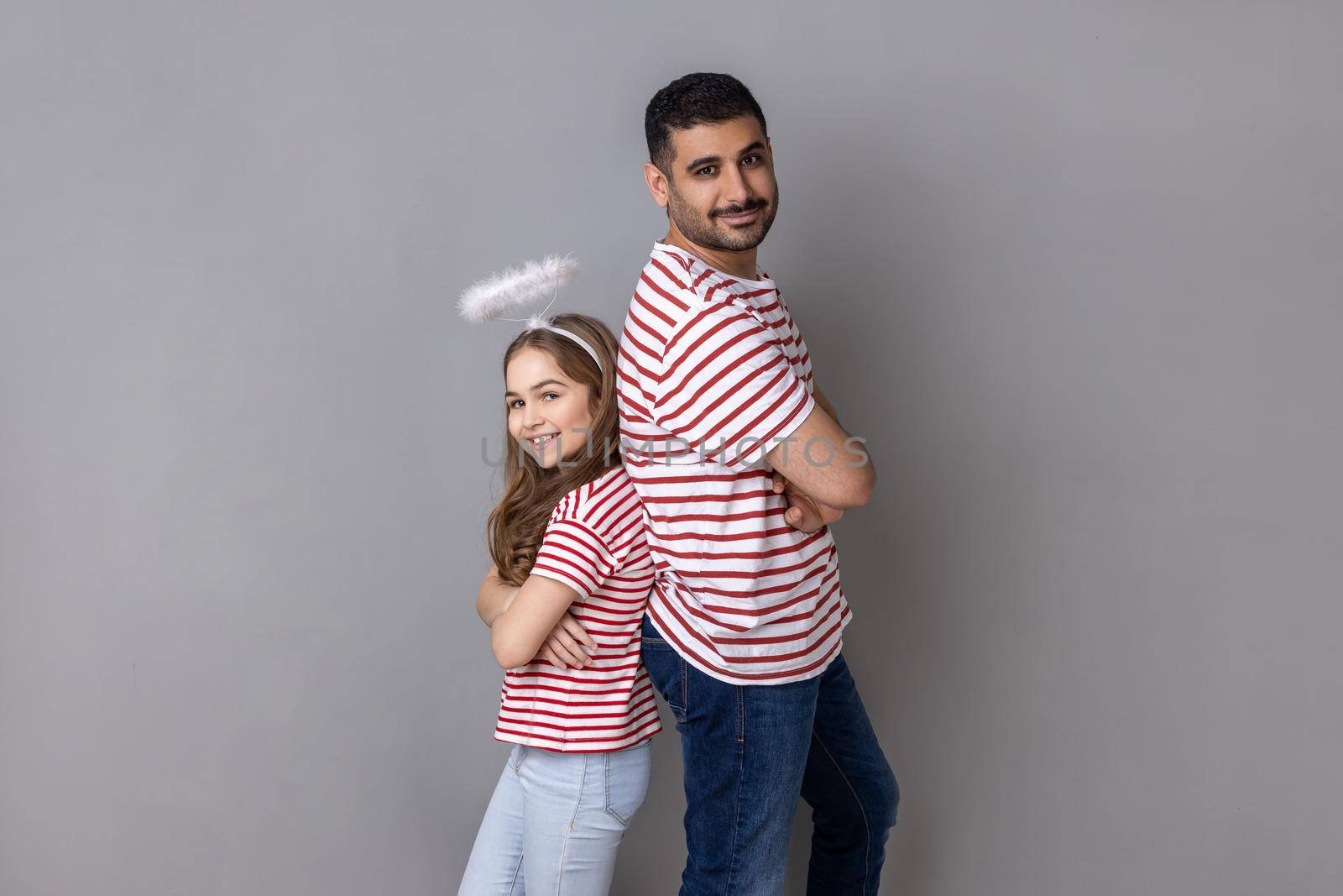 Portrait of lovely father and daughter in striped T-shirts, man and child with halo above head standing back to back with folded hands. Indoor studio shot isolated on gray background.