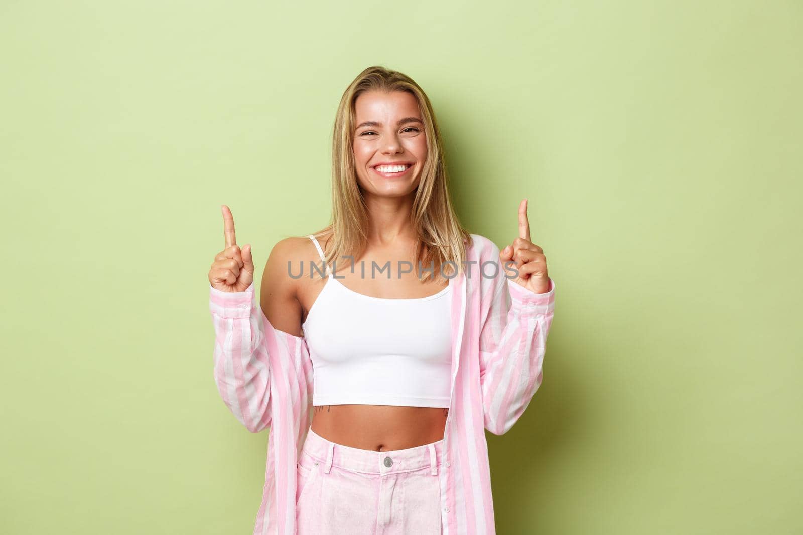 Image of cheerful smiling girl with short blond hairstyle, showing your logo or advertisement on green background, pointing fingers up.