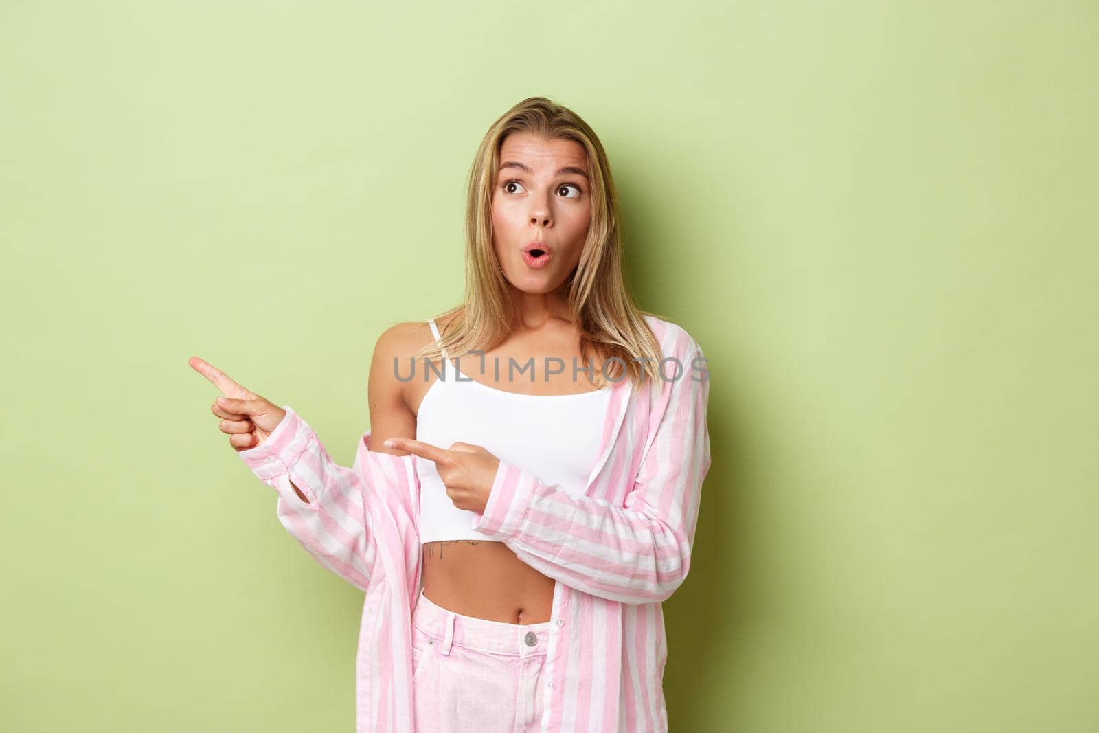 Portrait of beautiful blond girl in pink outfit, looking impressed and pointing fingers left, showing logo or product, standing over green background by Benzoix