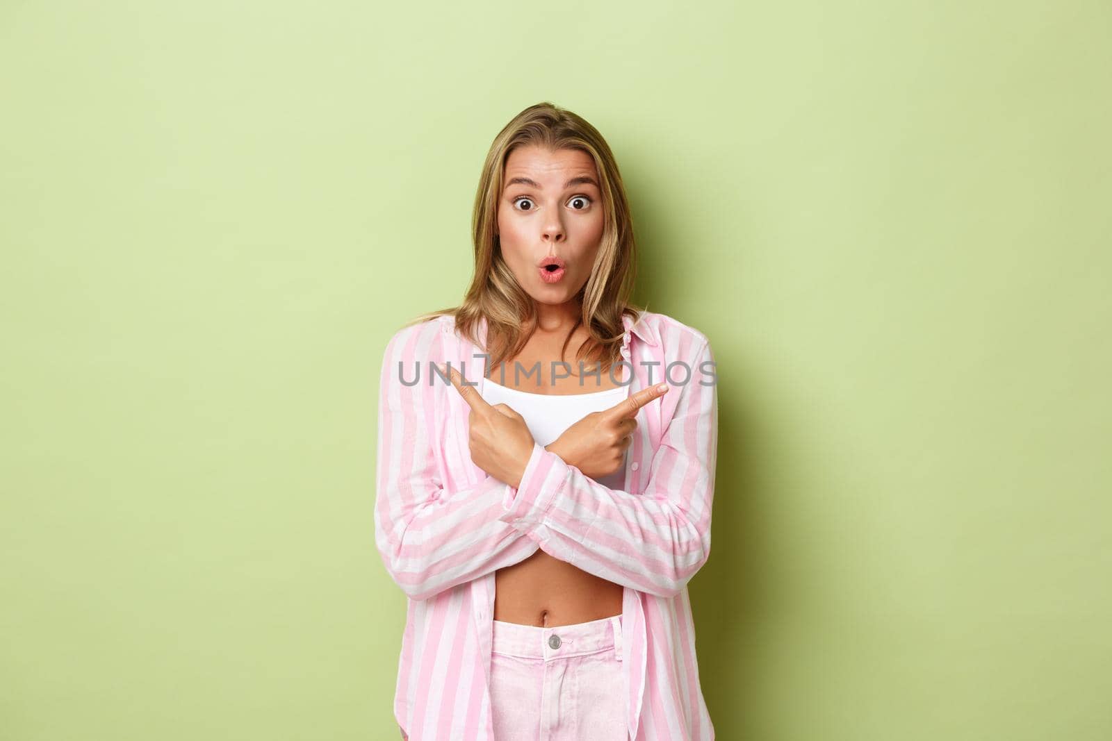 Portrait of surprised blond trendy girl in pink shirt with jeans, gasping amazed, pointing fingers sideways at two awesome choices, standing over green background.