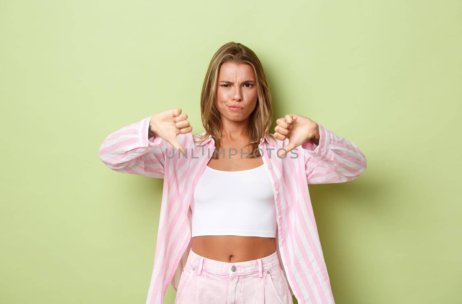 Image of disappointed blond woman in stylish pink outfit, frowning and showing thumbs-down, judging bad product, standing over green background by Benzoix