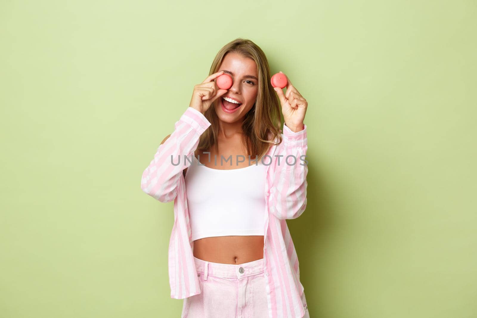 Portrait of beautiful modern woman with blond hair, showing pink macaroons and smiling, standing happy over green background.