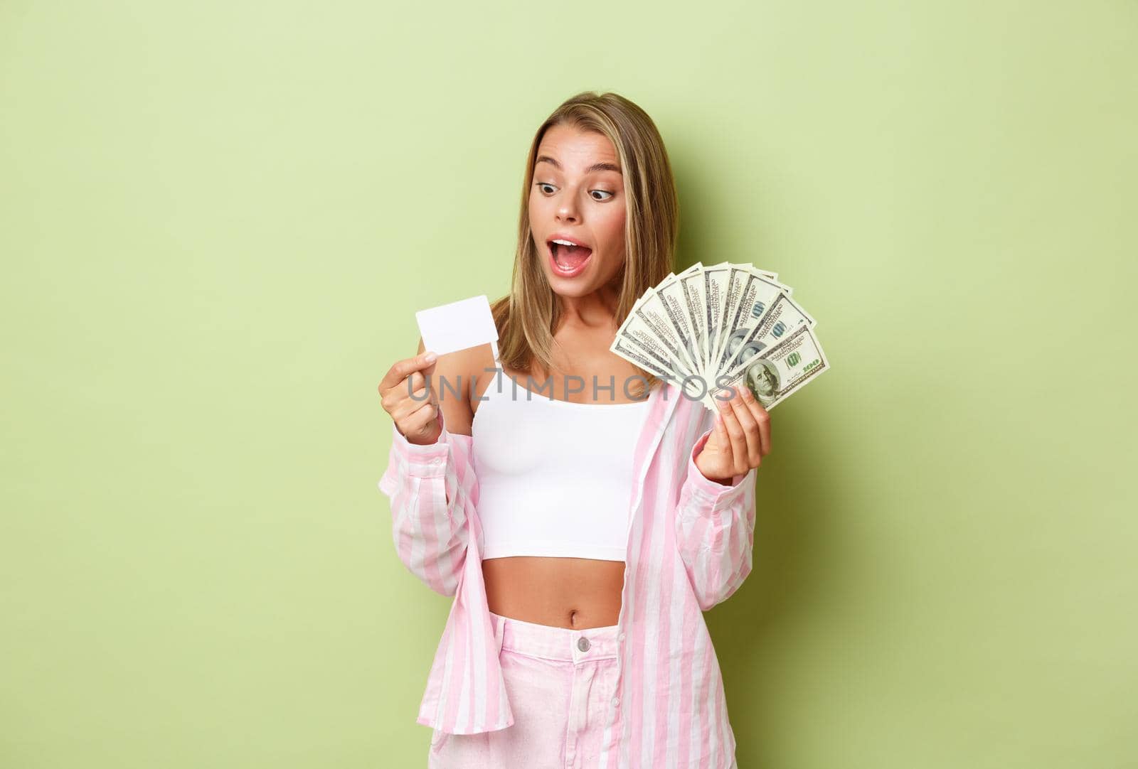 Portrait of attractive rich woman with blond hair, holding money and credit card and smiling amazed, standing over green background by Benzoix