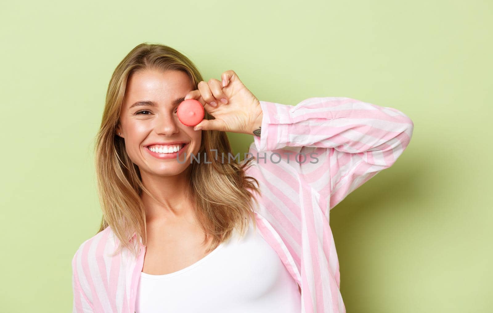 Close-up of playful smiling blond girl, holding macaroon near eye, standing happy over green background.