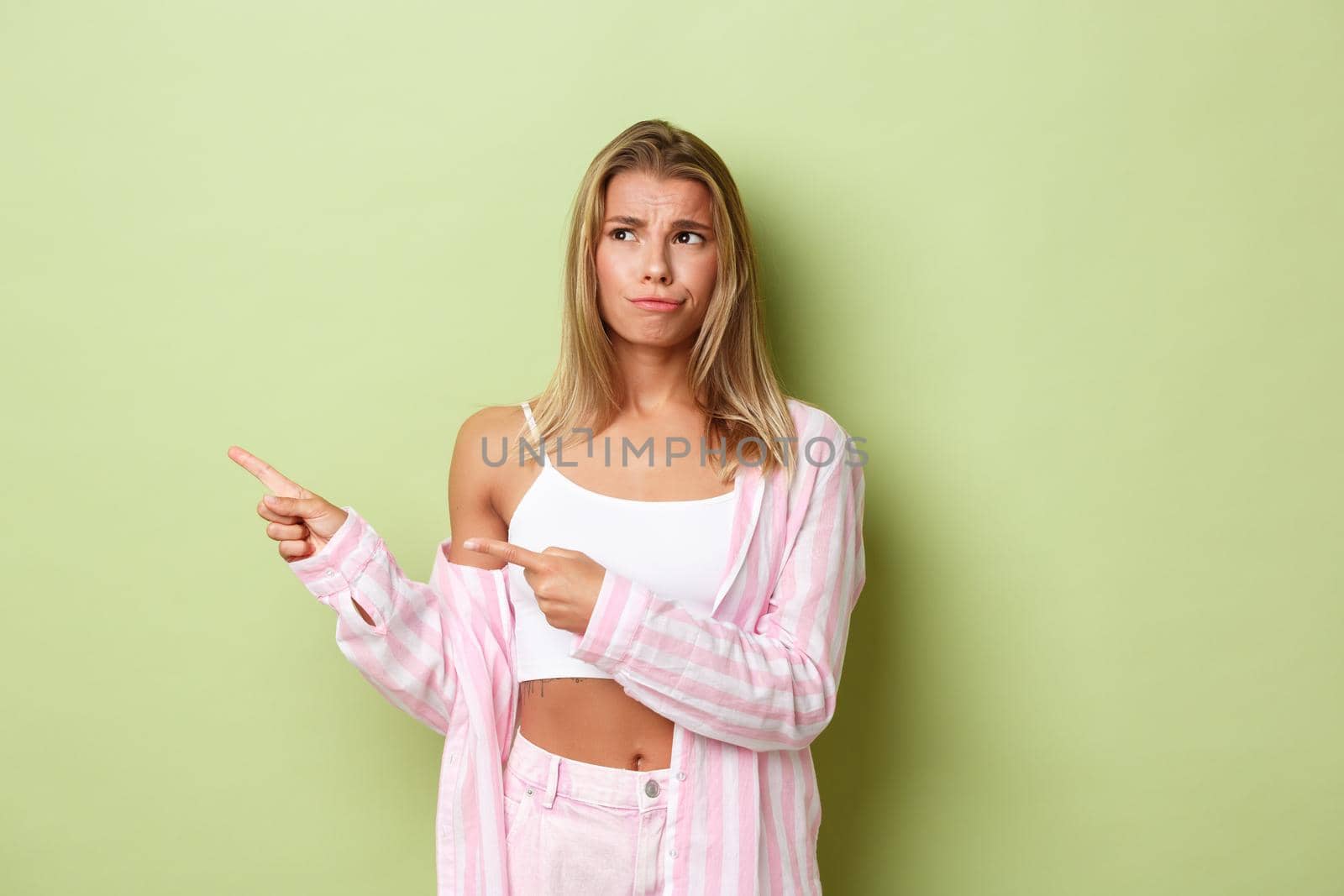 Skeptical and disappointed blond female in pink costume looking doubtful, frowning and pointing fingers left at logo, standing over green background.