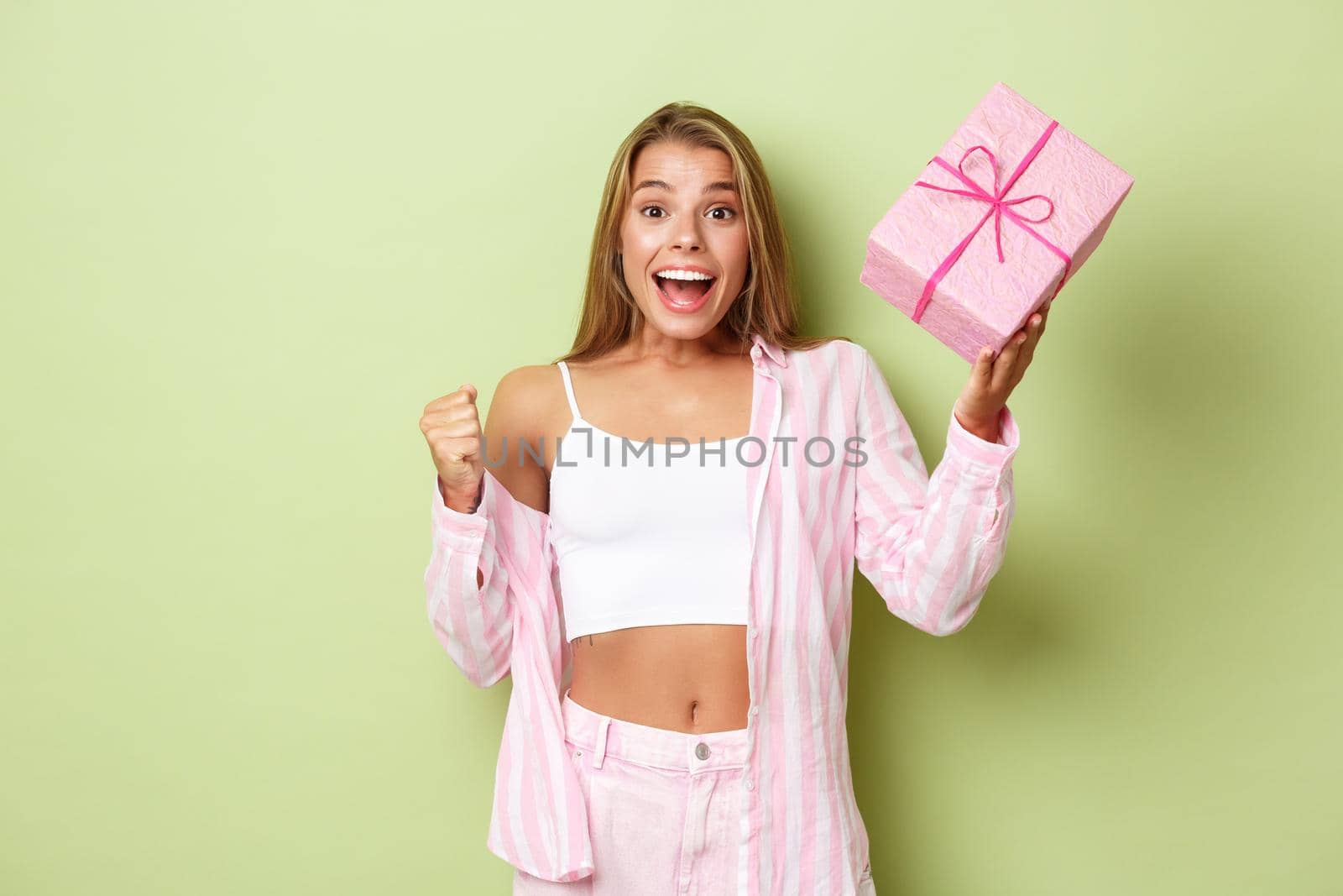 Cheerful blond girl celebrating holiday, holding gift and looking excited, standing over green background.