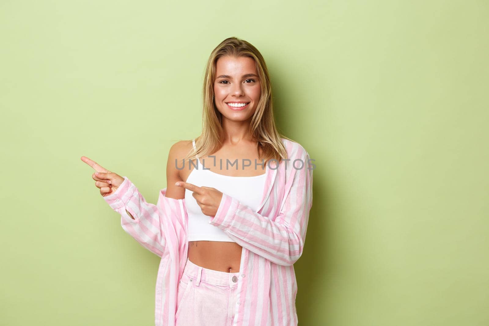 Attractive smiling blond girl recommending promo, pointing fingers left at your logo on green empty space, standing in stylish pink outfit.