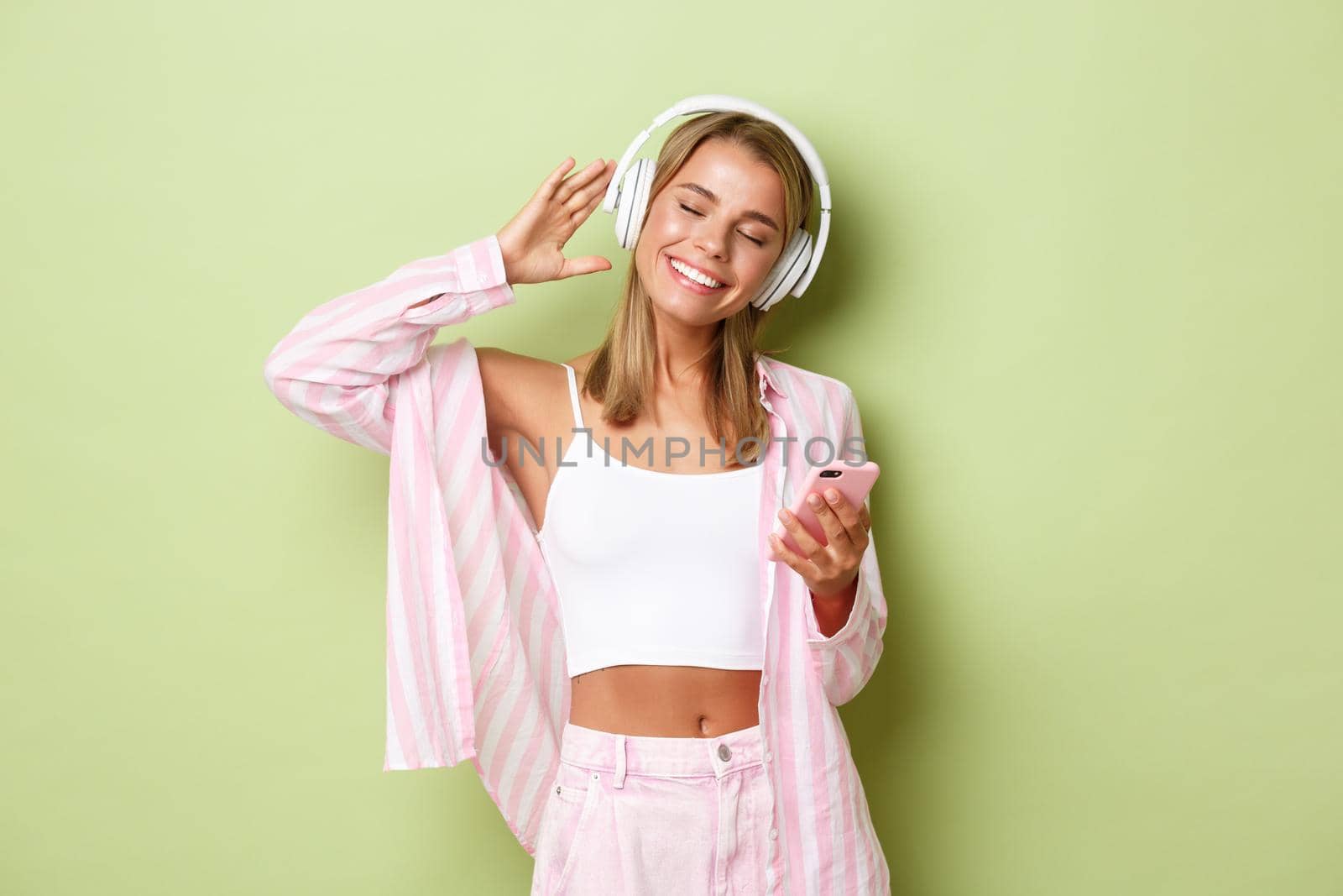Portrait of attractive happy woman with blond short hair, wearing pink shirt, listening music in wireless headphones and smiling, holding mobile phone, standing over green background.
