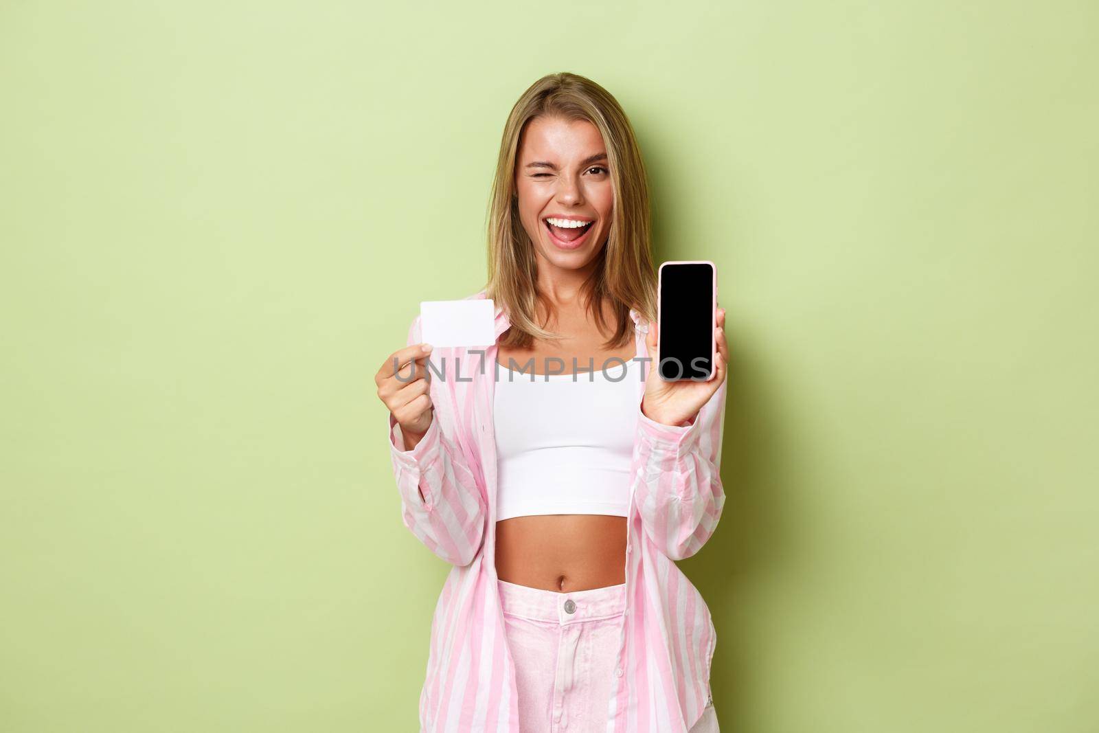 Image of sassy modern girl with blond short hair, showing credit card and mobile phone screen, winking and smiling while advertising application, standing over green background.