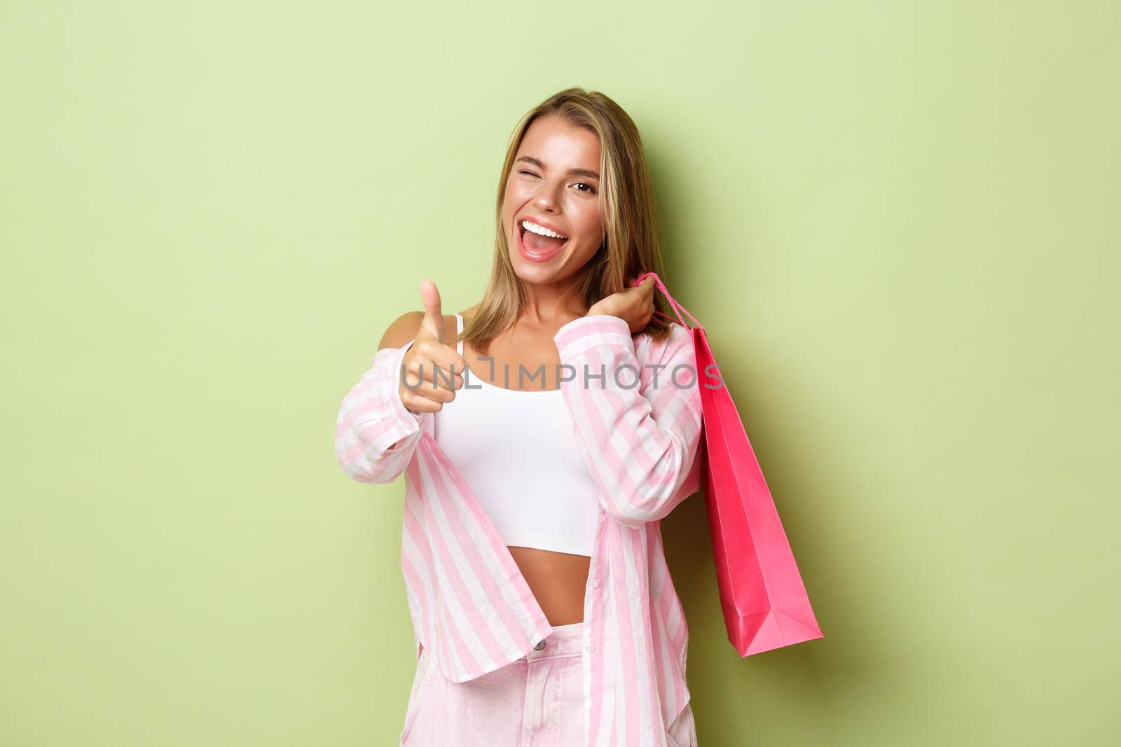 Portrait of attractive glamour girl smiling, showing thumbs-up and holding shopping bag, winking at camera, standing over green background by Benzoix