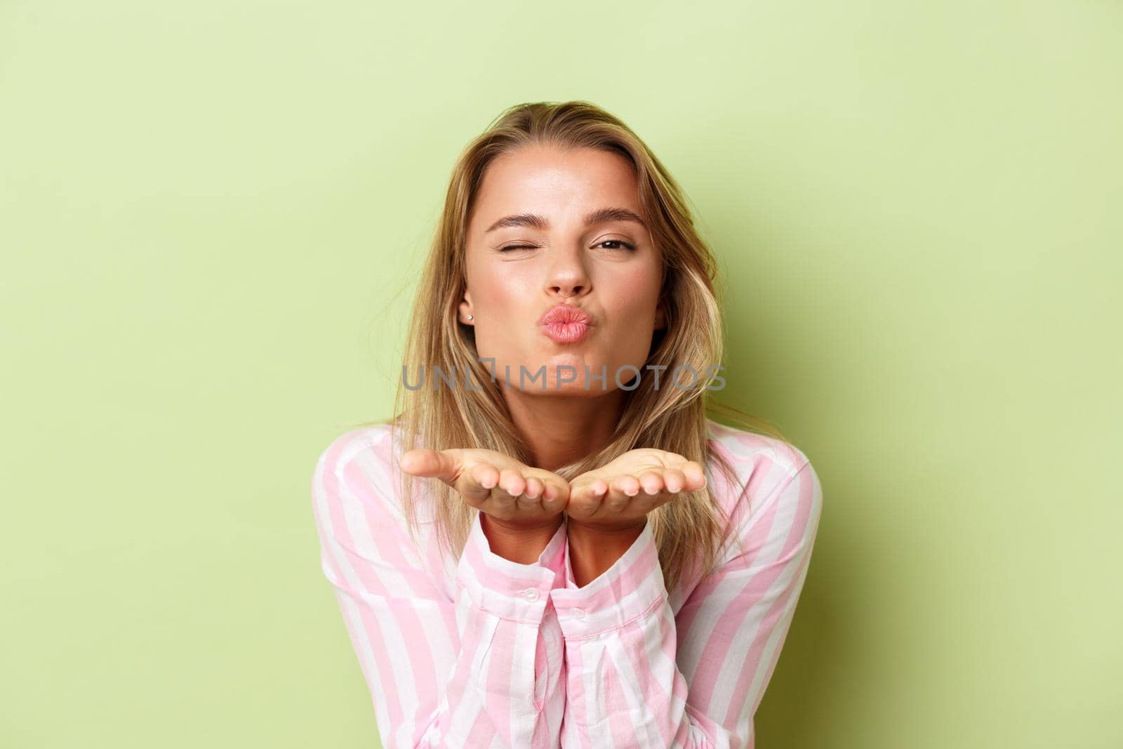 Close-up of beautiful blond girl in pink shirt, sending air kiss at camera with lovely expression, standing over green background.