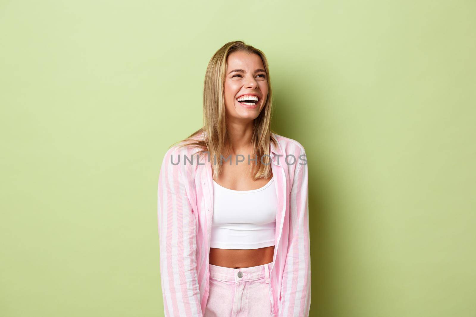 Portrait of happy beautiful woman with short blond hair, starting to laugh and looking at upper right corner, smiling happy, standing in pink shirt and jeans over green background.